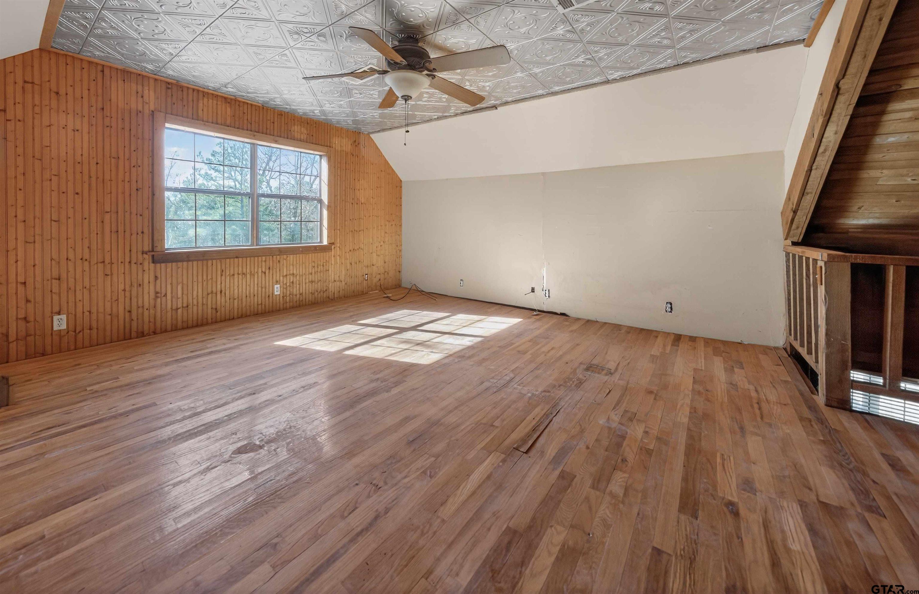 Empty room with hardwood floors, wood-paneled walls, and a large sunlit window.