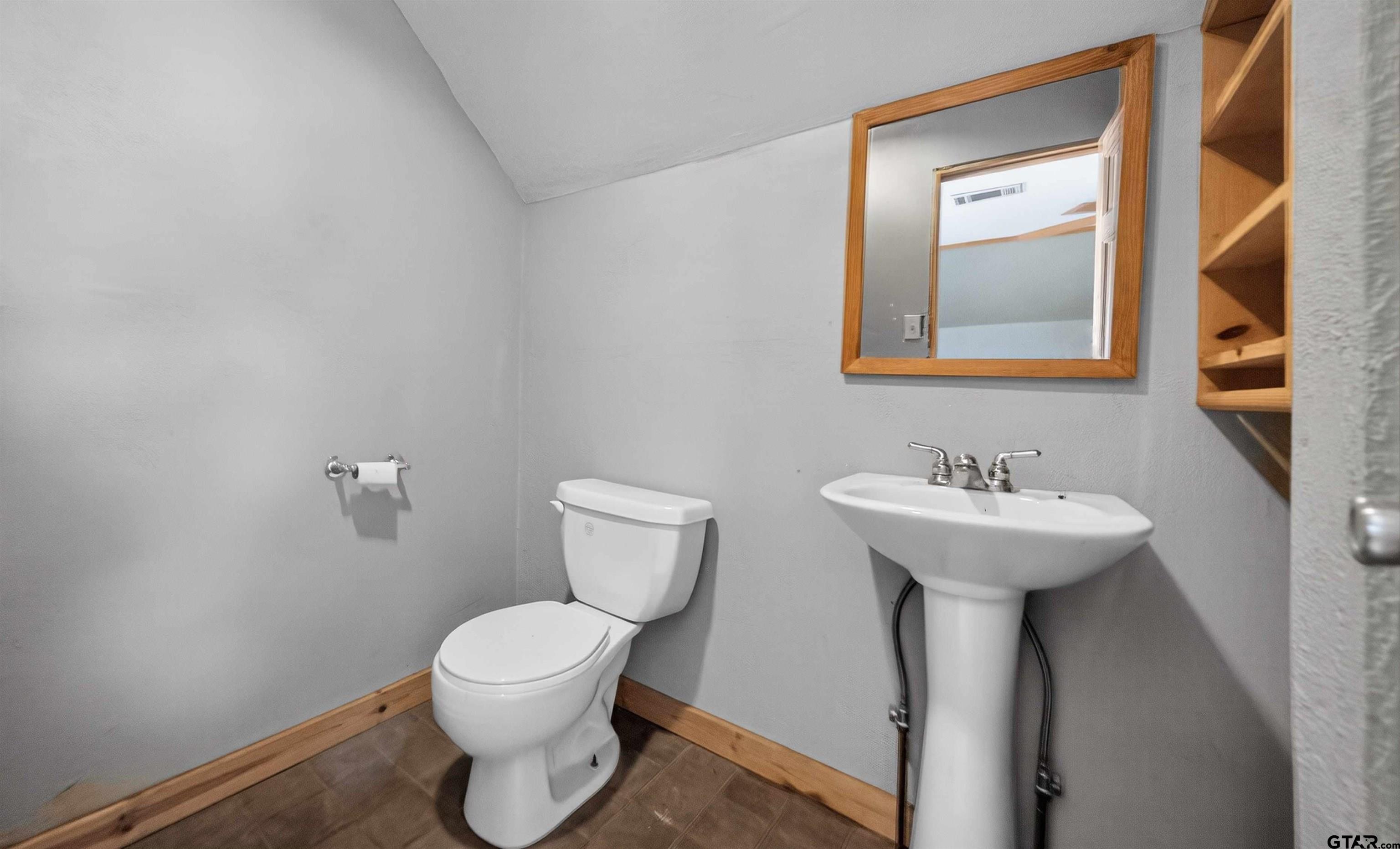Bathroom with a white toilet, pedestal sink, wood-framed mirror, and open wooden shelving.