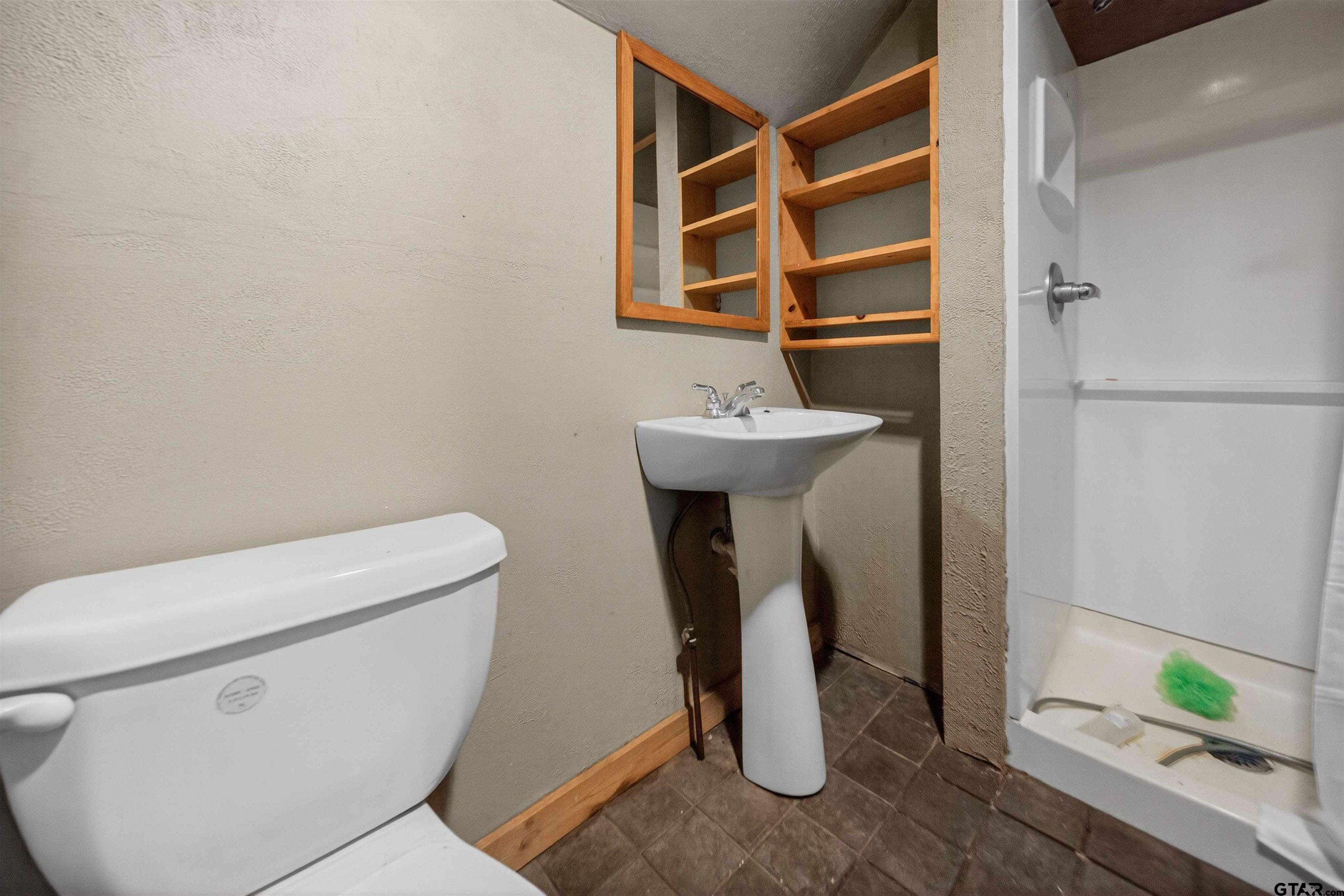 Bathroom with pedestal sink, open wooden cabinet, toilet, and shower stall.