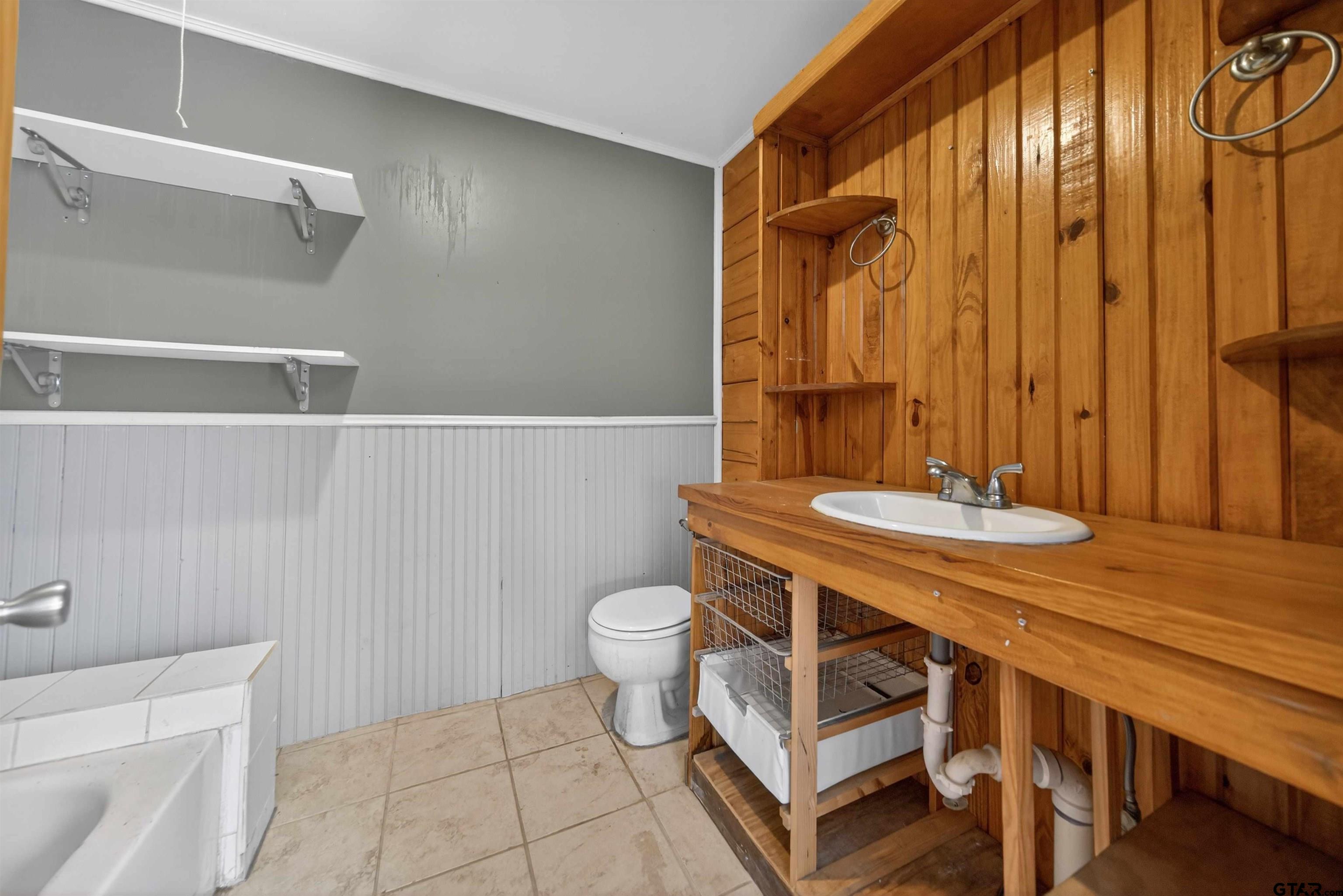 Bathroom with a rustic wood vanity, inset sink, exposed plumbing, white toilet, and tiled floor.