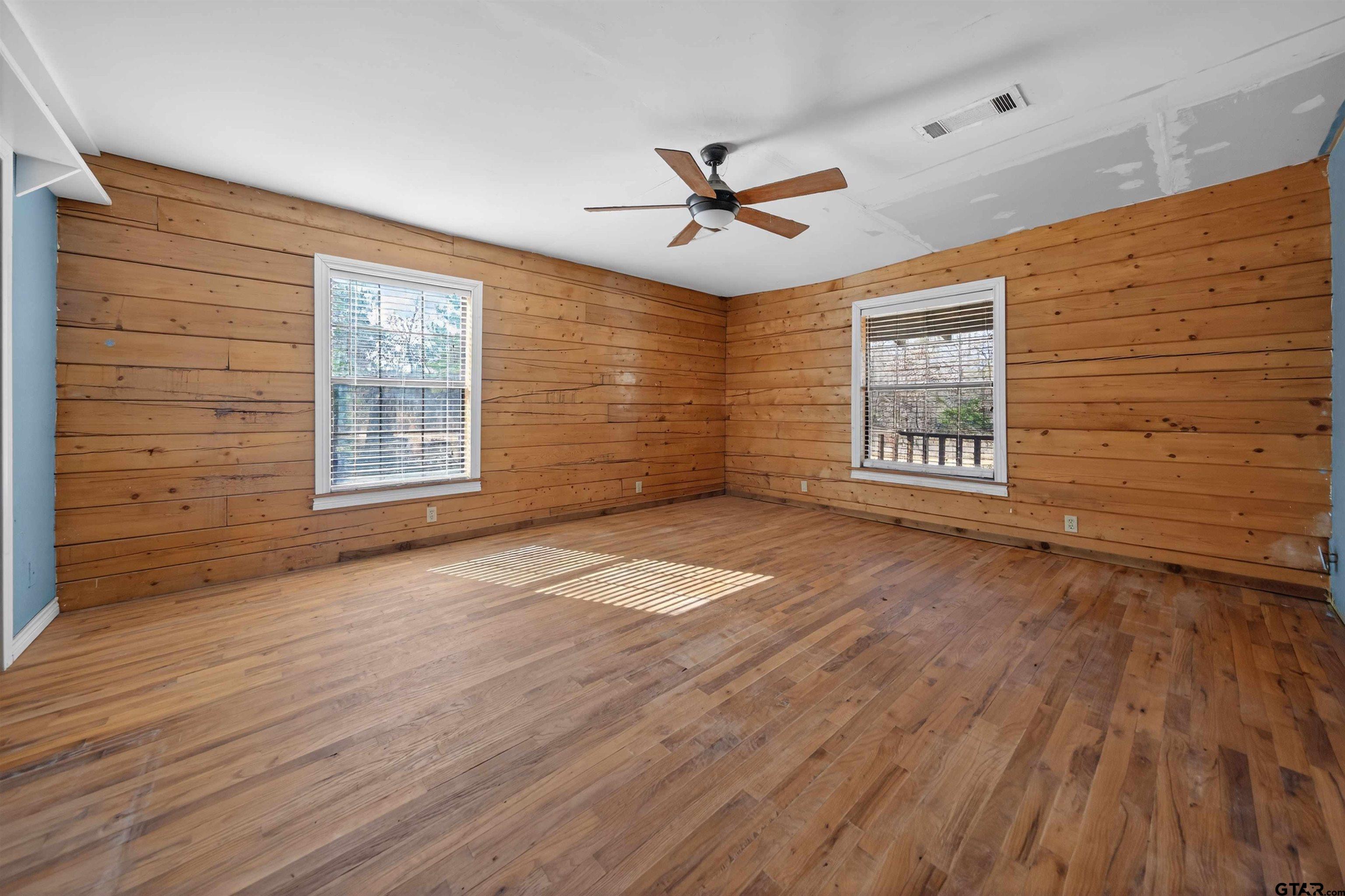 Empty room with wood-plank walls, hardwood floors, two windows, and a ceiling fan.