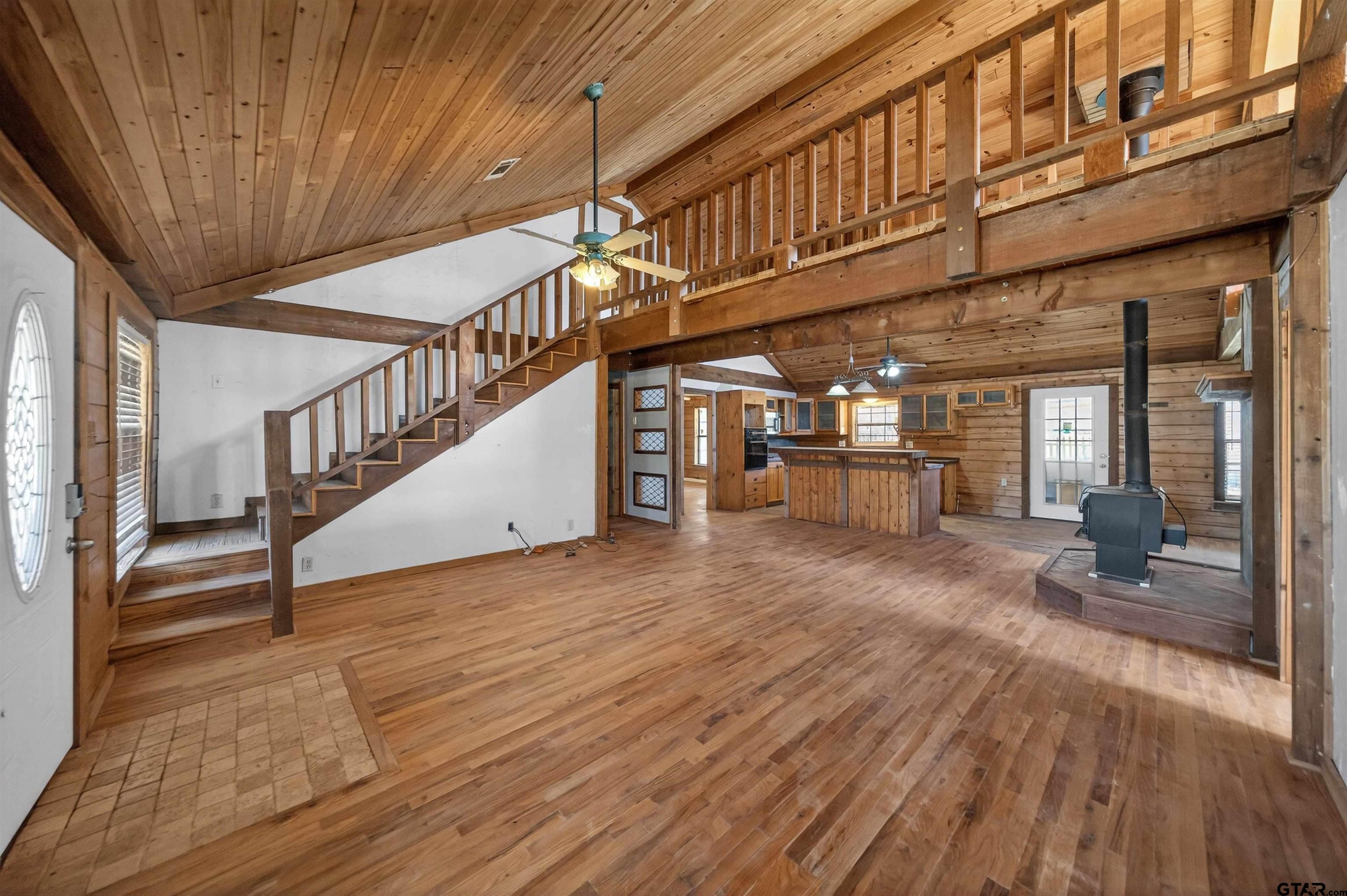 Open living room with wood floors, staircase to a loft, kitchen island, and wood-burning stove.