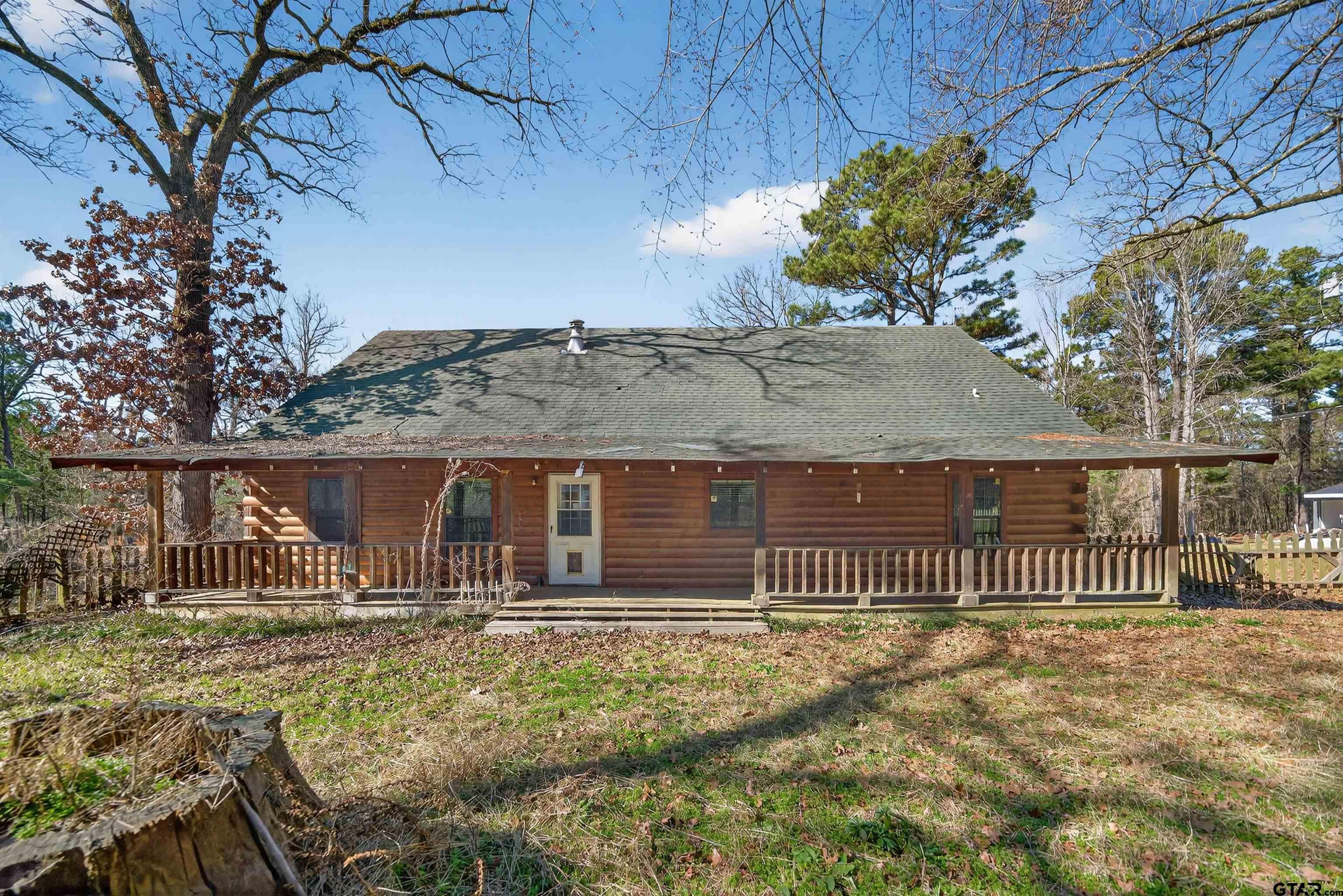 Log cabin with a long covered porch, gray shingle roof, and wooded yard.