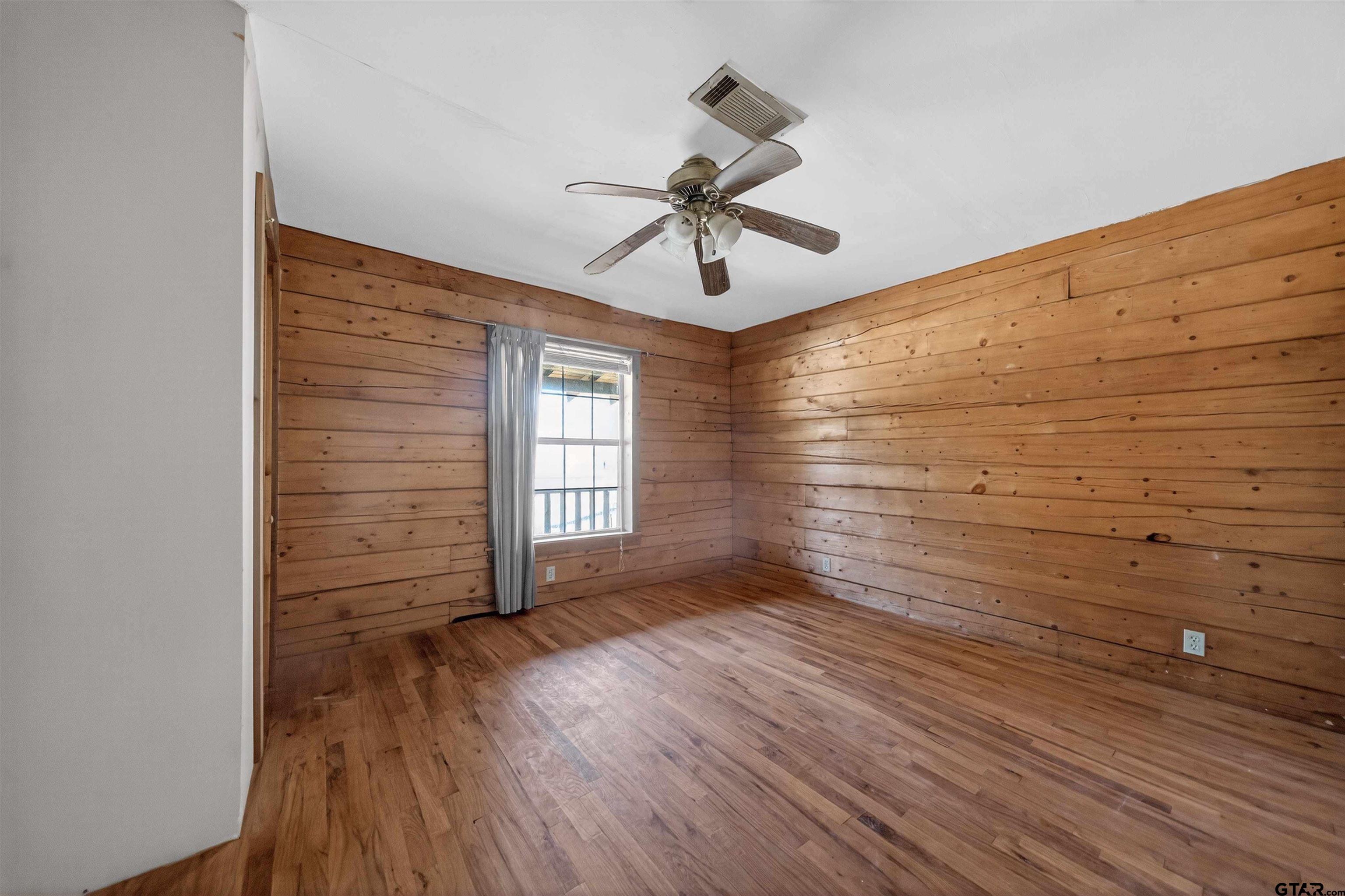 Empty room with wood-paneled walls, hardwood flooring, a curtained window, and ceiling fan.