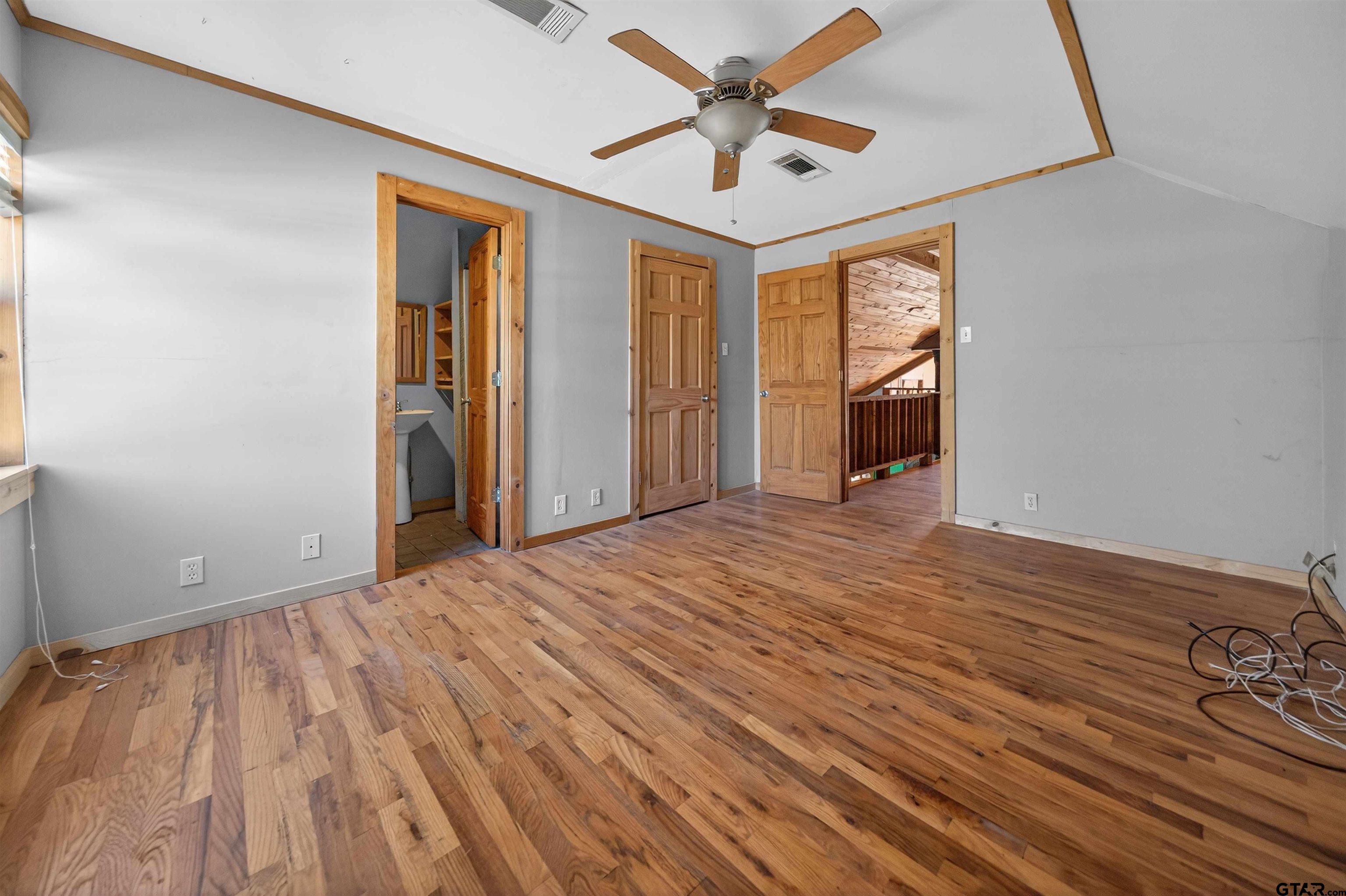 Empty bedroom with hardwood floors, a wood-trimmed ceiling fan, and open doorways to a bathroom and hall.