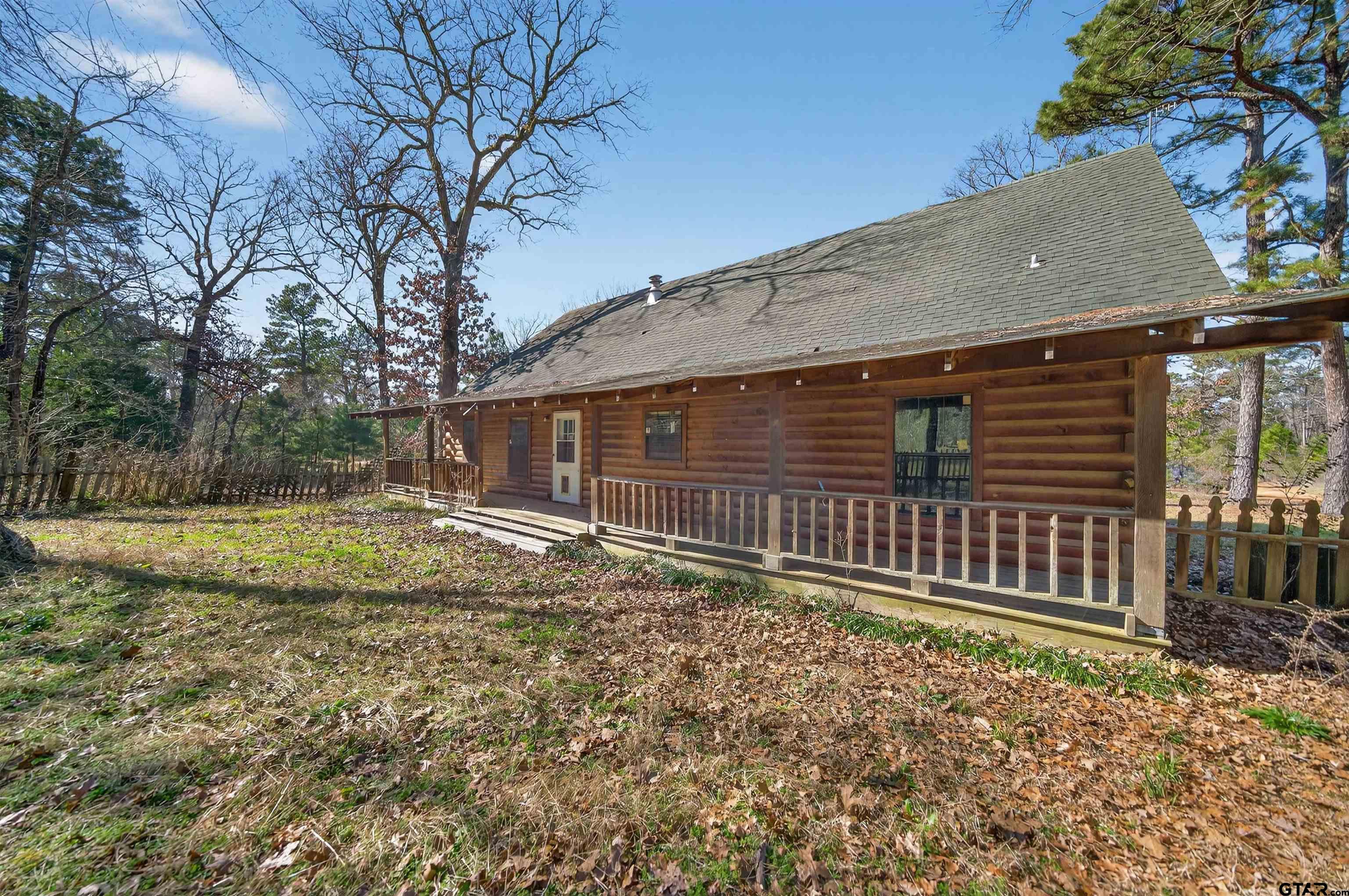 Log cabin with a long covered porch among trees and fallen autumn leaves.