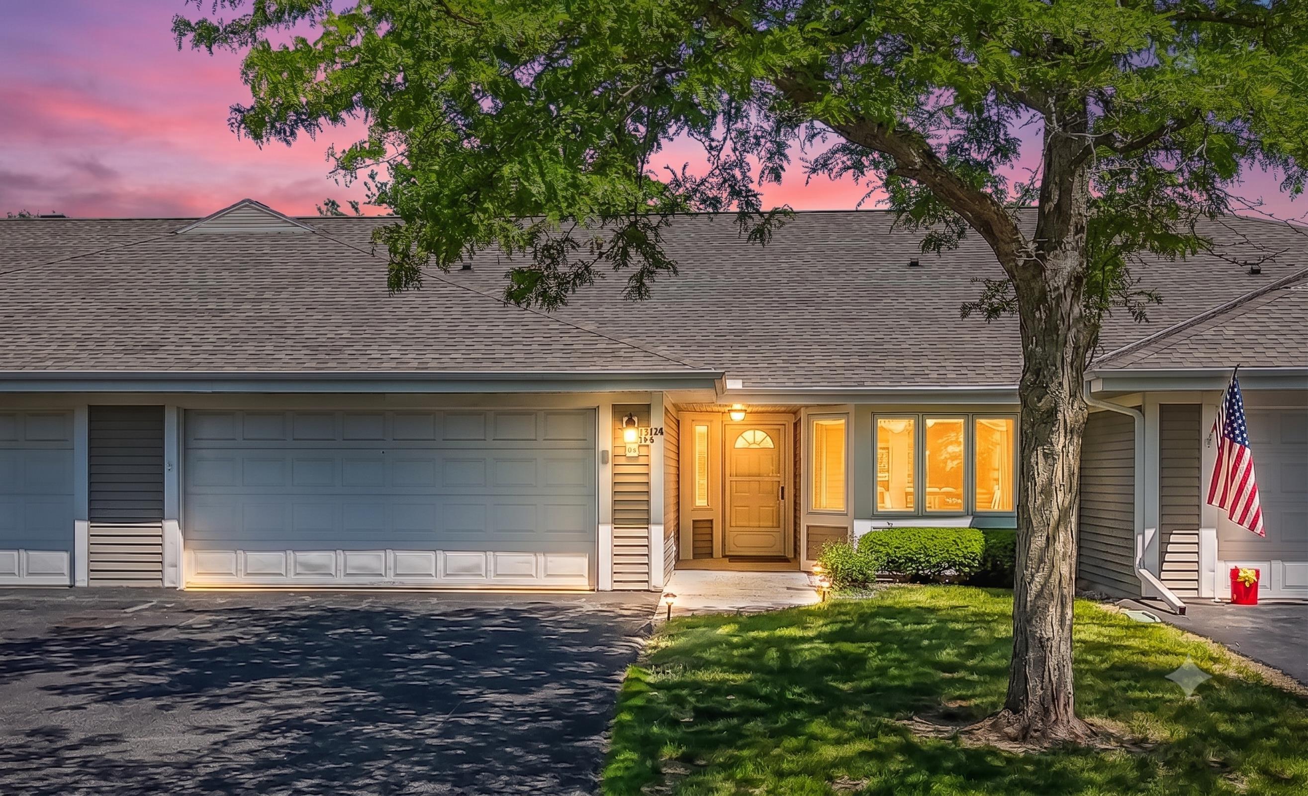 Gray single-story home with a double garage, warmly lit entry, large shade tree, and American flag at dusk.