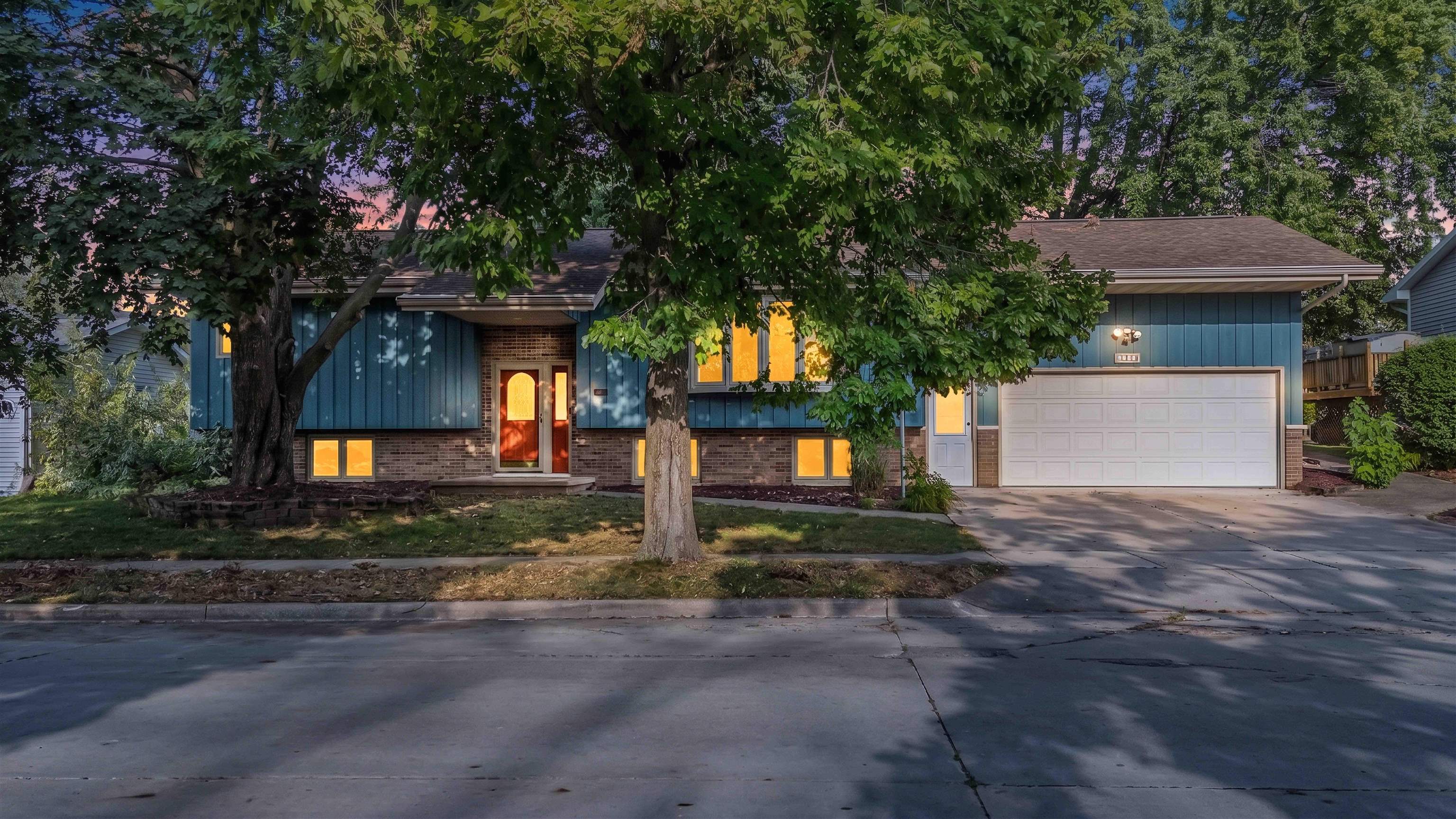 Blue split-level home with brick accents and an attached garage beneath mature trees at dusk.