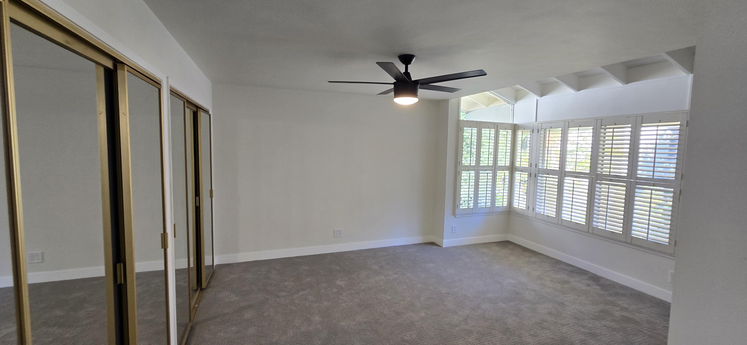 Empty bedroom with mirrored closet doors, gray carpet, and plantation-shuttered corner windows.