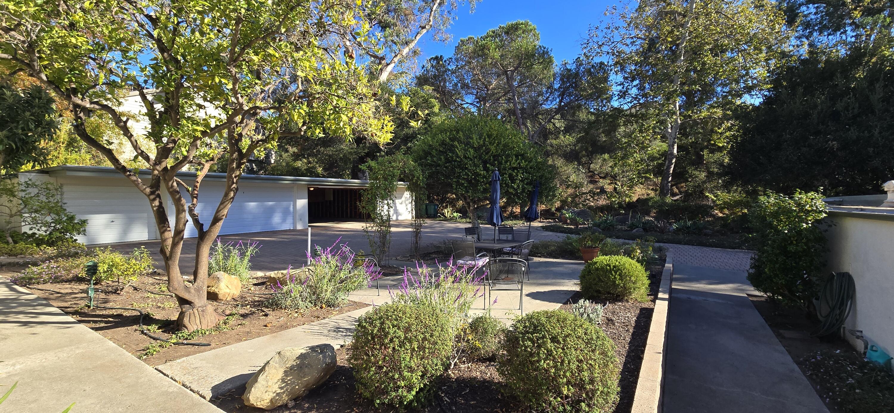 Landscaped courtyard with patio seating, flowering plants, mature trees, and a white garage.