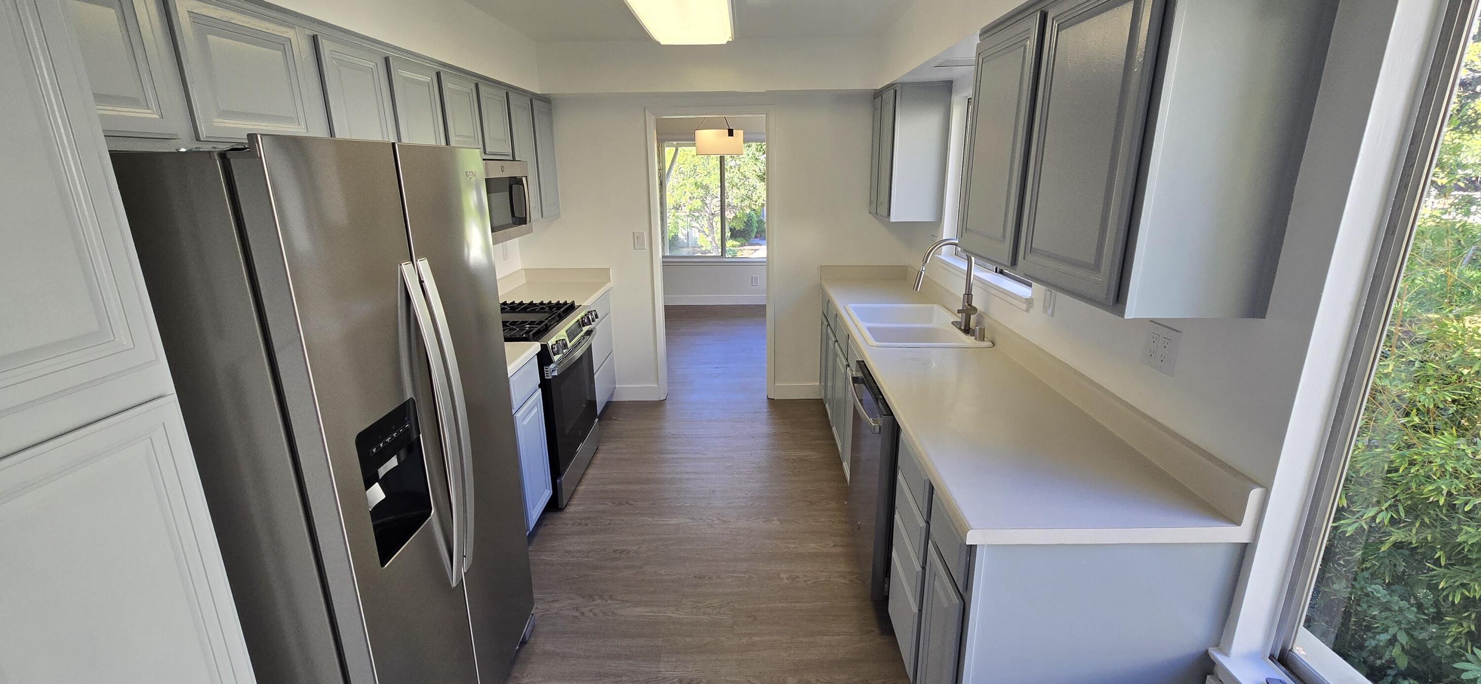 Galley kitchen with gray cabinets, stainless steel appliances, white counters, and windows overlooking greenery.
