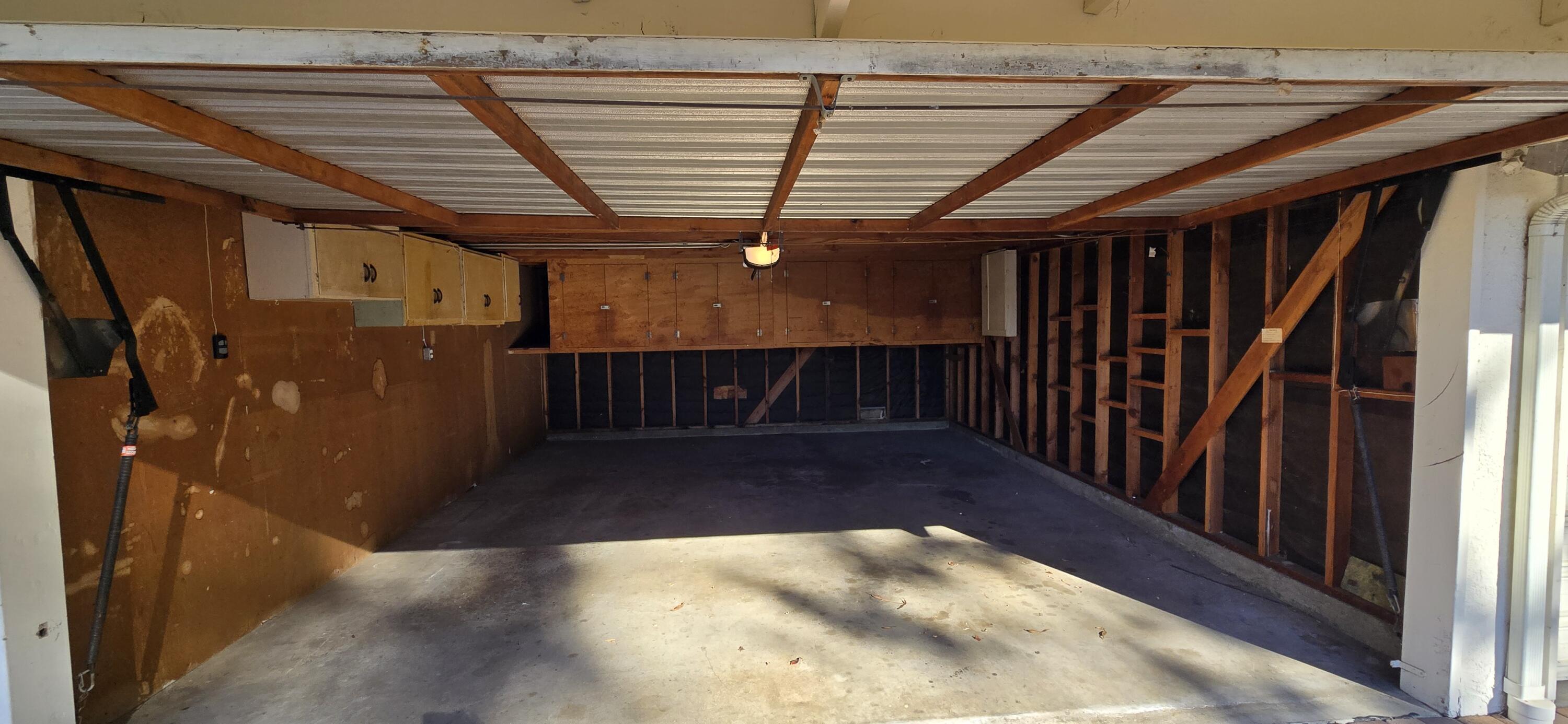 Empty garage with exposed wooden framing, wall cabinets, and sunlight across the concrete floor.