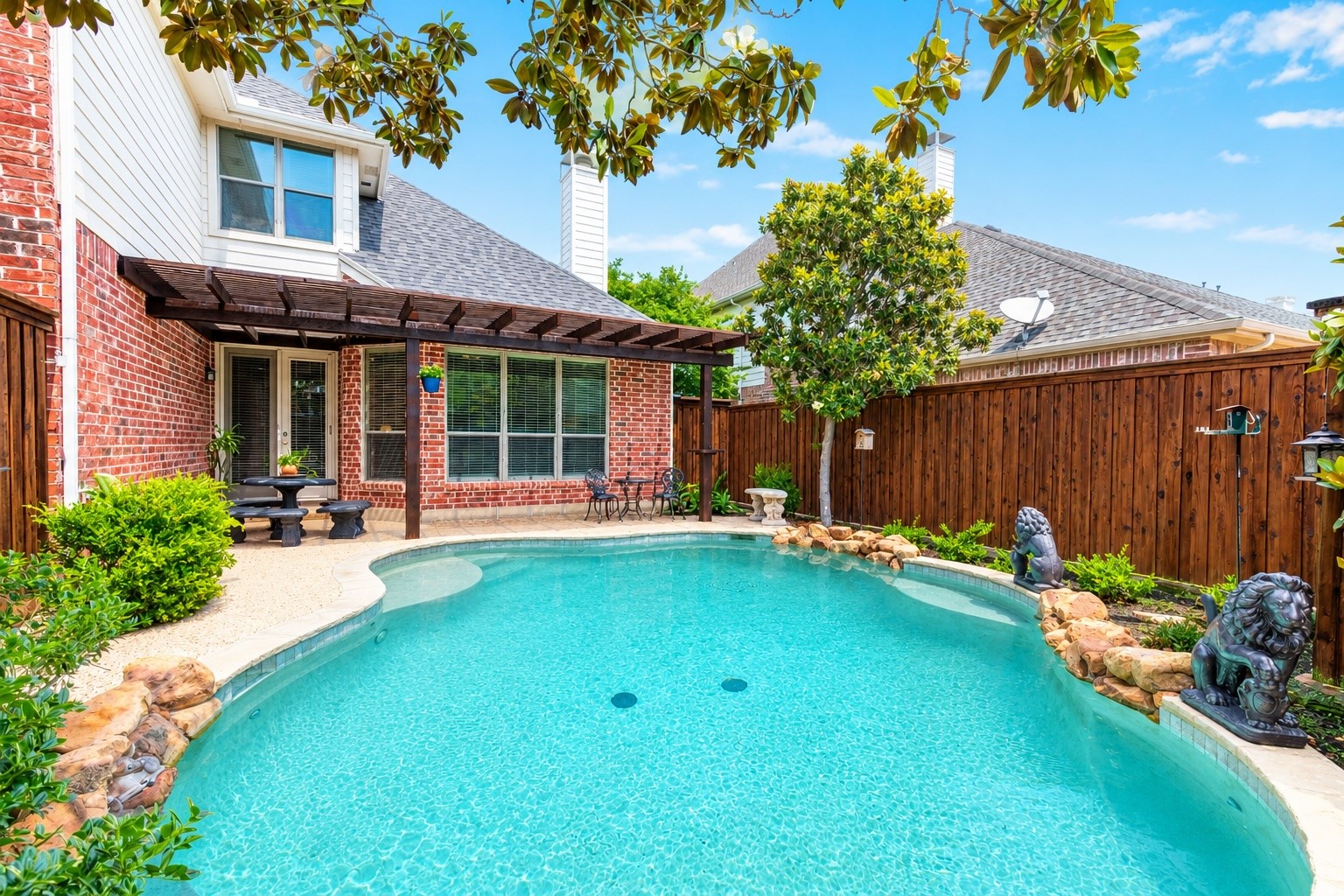 Backyard swimming pool beside a brick home, with a wooden pergola and fenced patio.