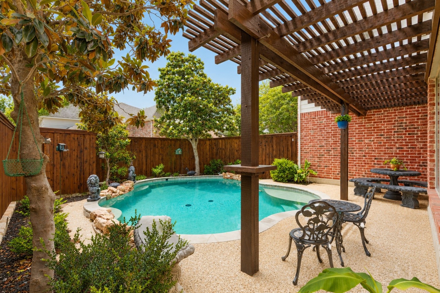 Backyard pool beneath a wooden pergola, with stone edging and patio dining furniture.