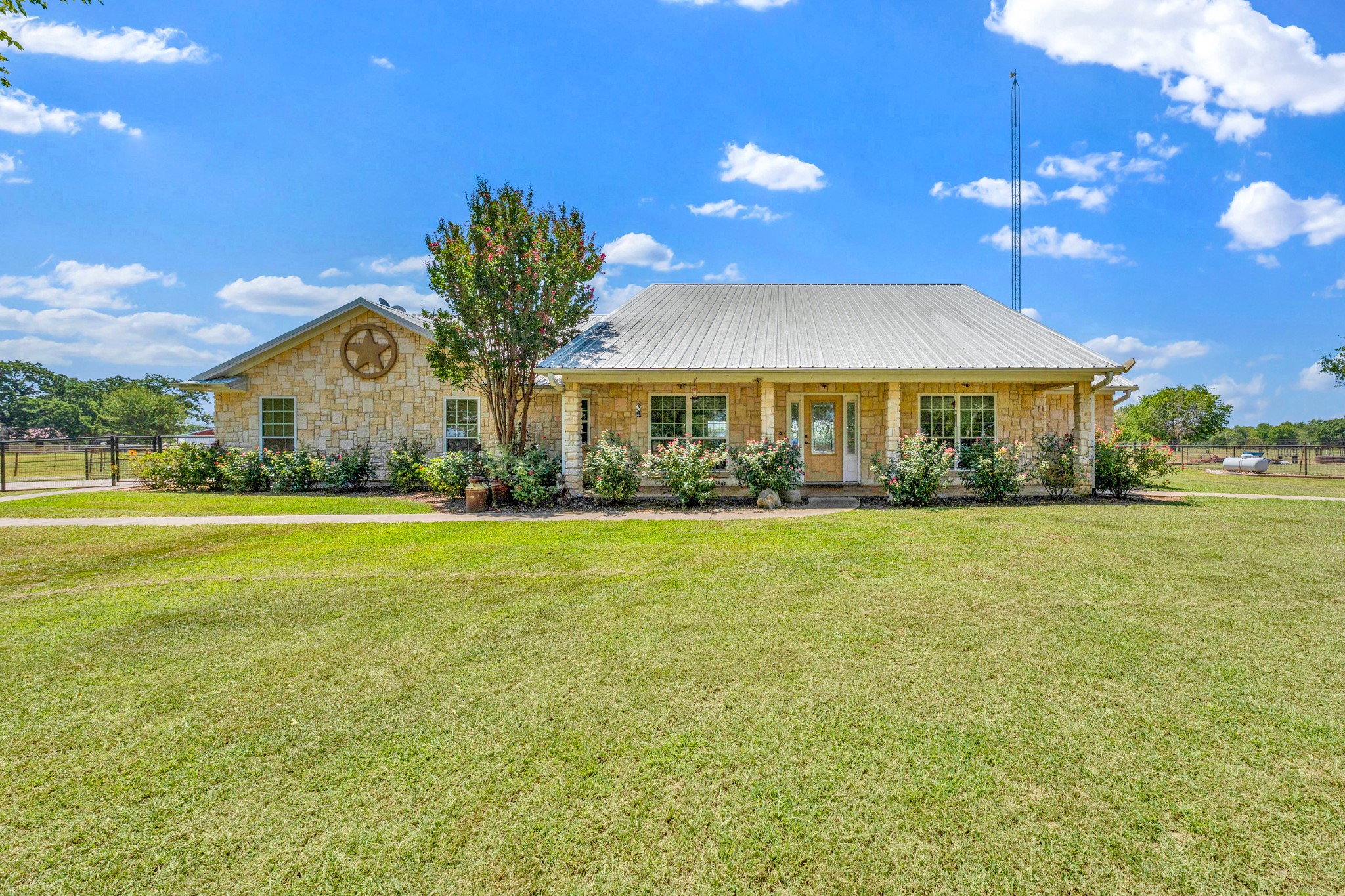 Stone house with a metal roof, star emblem, flowering shrubs, and broad lawn.