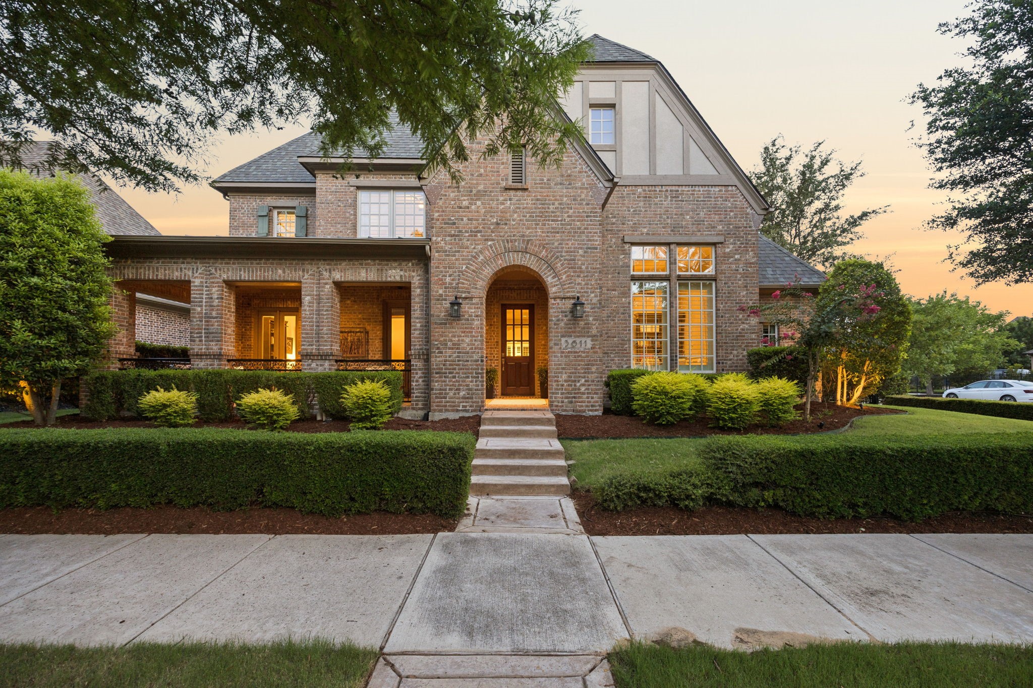 Two-story brick house with an arched entry, covered porch, and trimmed hedges at dusk.