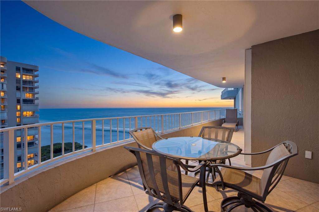 Furnished balcony overlooking the ocean at sunset, with a glass dining table and neighboring high-rise.