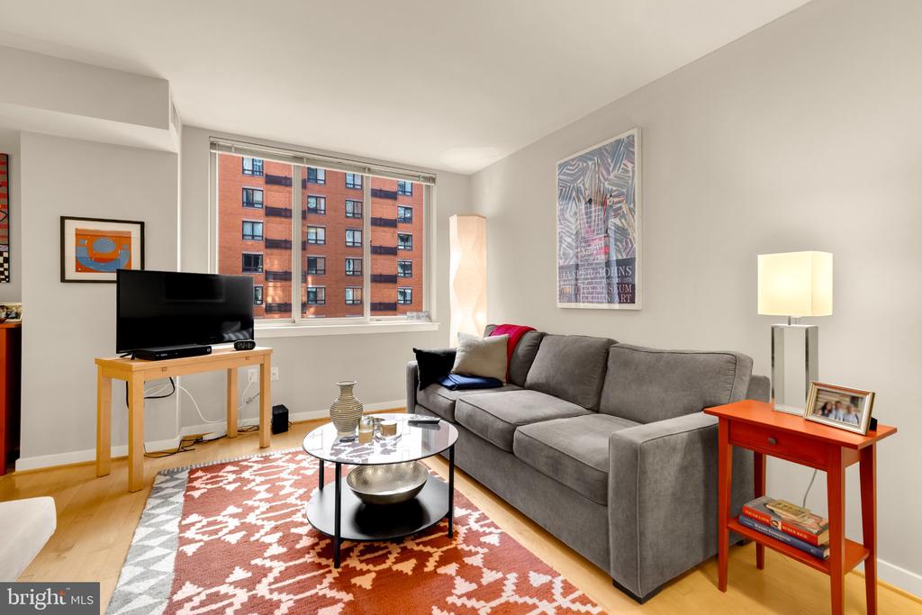Living room with a gray sectional, patterned rug, and large windows overlooking a brick apartment building.