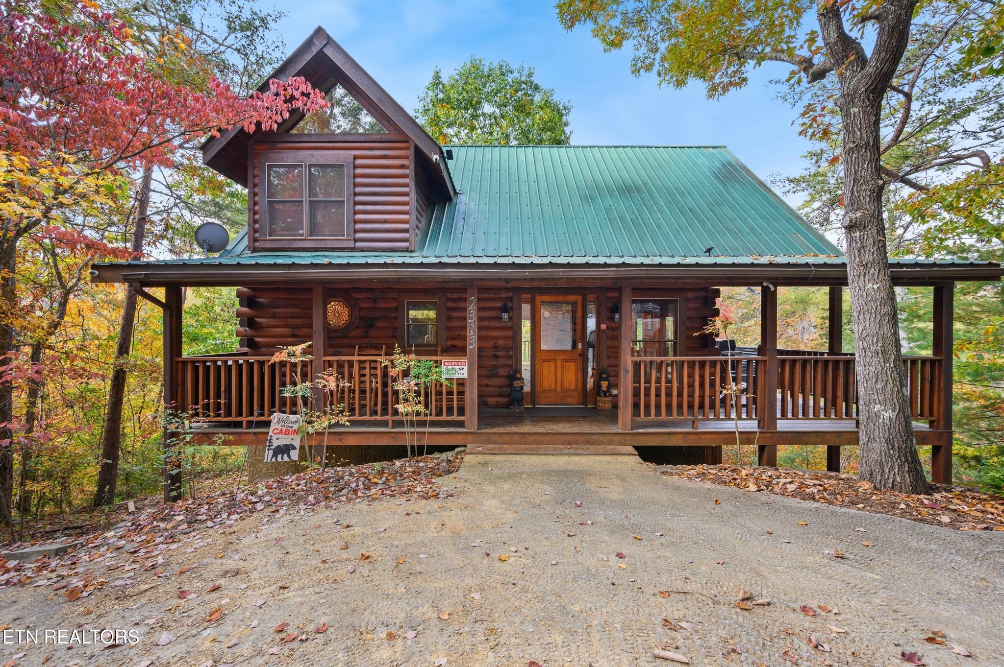 Two-story log cabin with a green metal roof and broad porch amid colorful autumn trees.