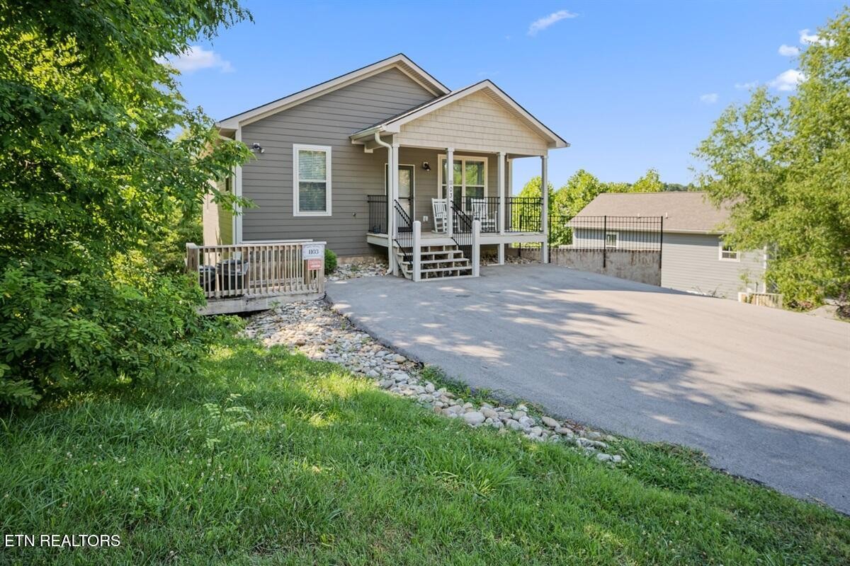 Gray-sided house with a covered porch and broad paved driveway, framed by leafy trees.
