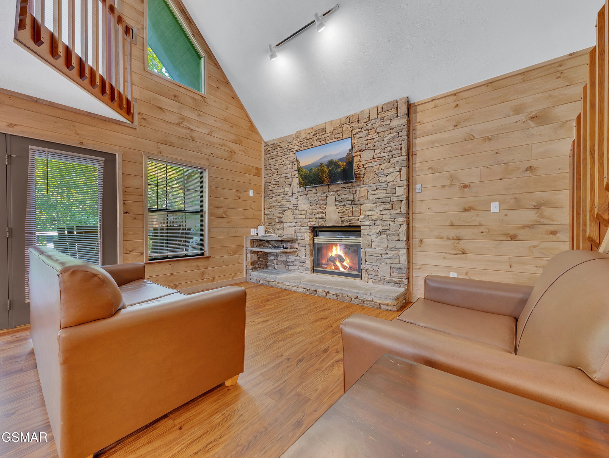 Cabin living room with a stone fireplace, mounted TV, vaulted wood walls, and brown leather seating.