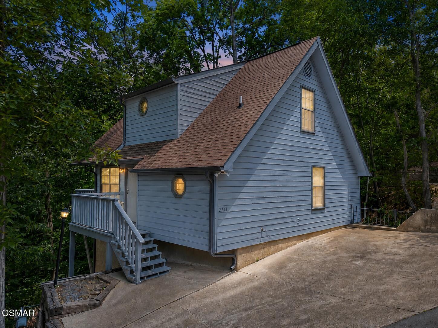 Blue-sided cabin with a steep gable roof, lit windows, entry stairs, and a paved driveway among trees.