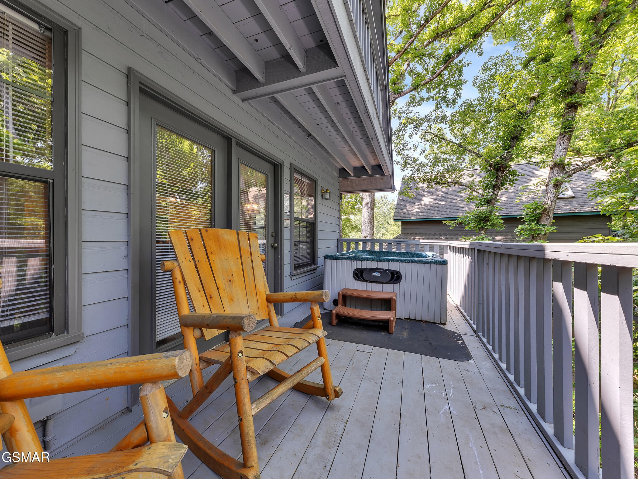 Covered deck with wooden chairs and a hot tub beside leafy trees.