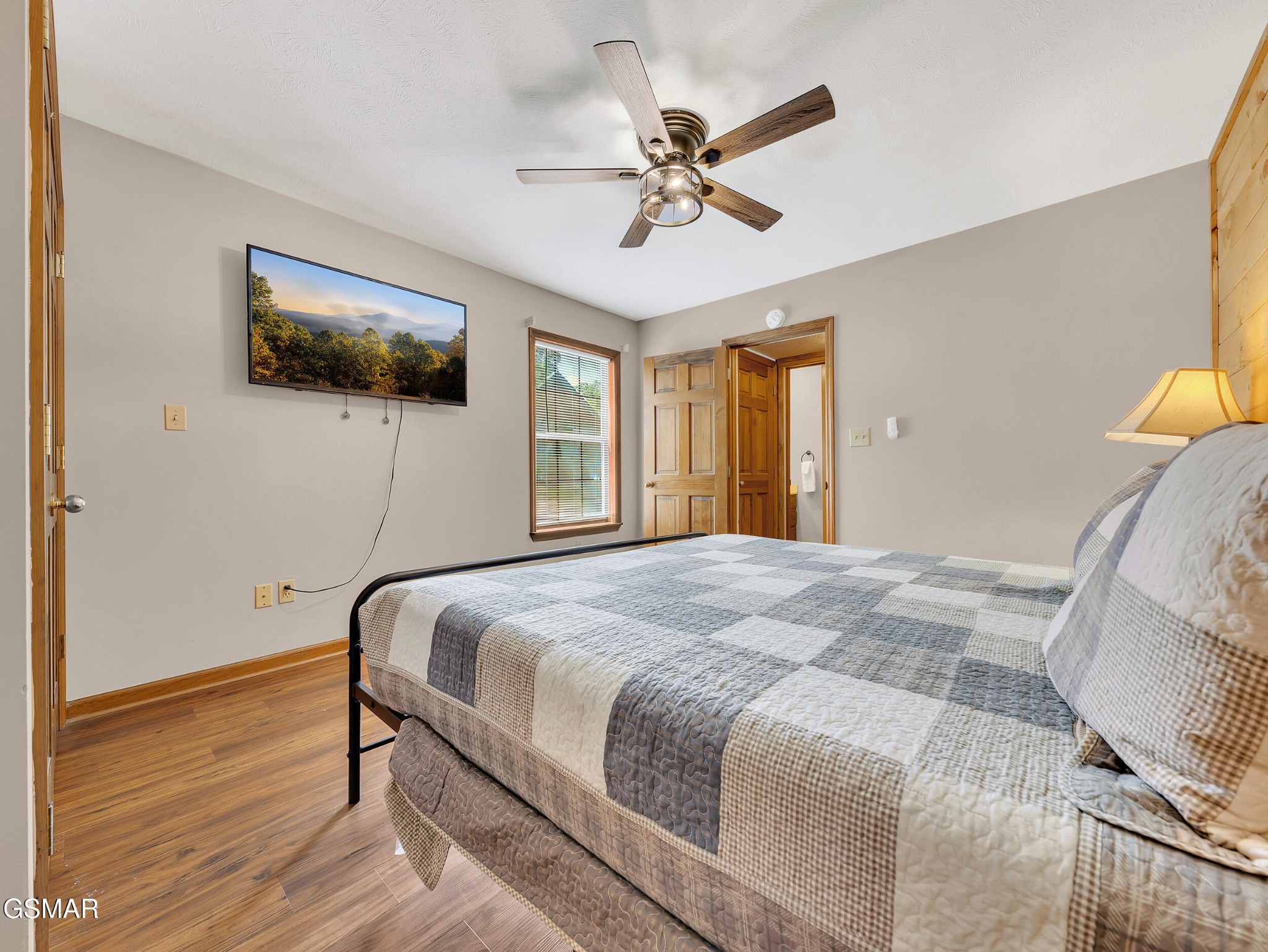 Bedroom with a patchwork-covered bed, wall-mounted TV, ceiling fan, and wood-look flooring.
