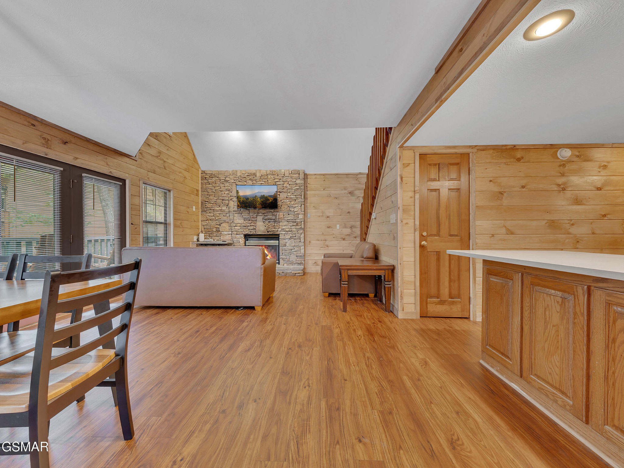 Open-plan living room with a stone fireplace, wood-paneled walls, and dining area by the windows.