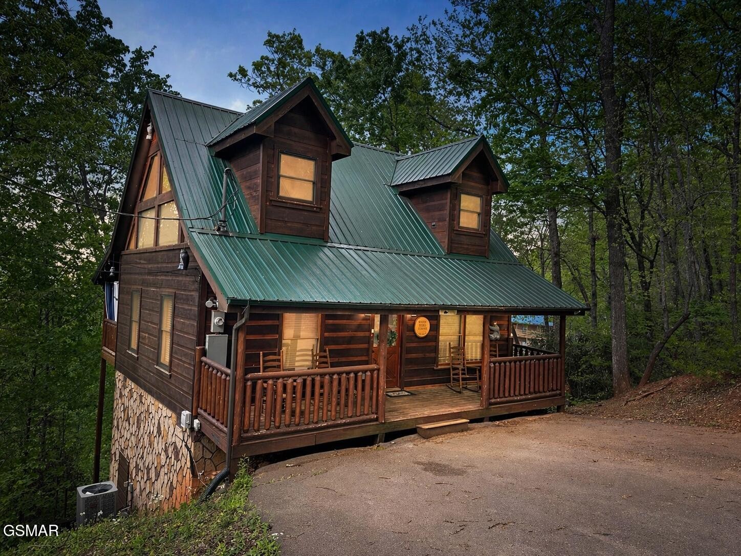 Wood-sided cabin with a green metal roof and covered porch nestled among trees at dusk.