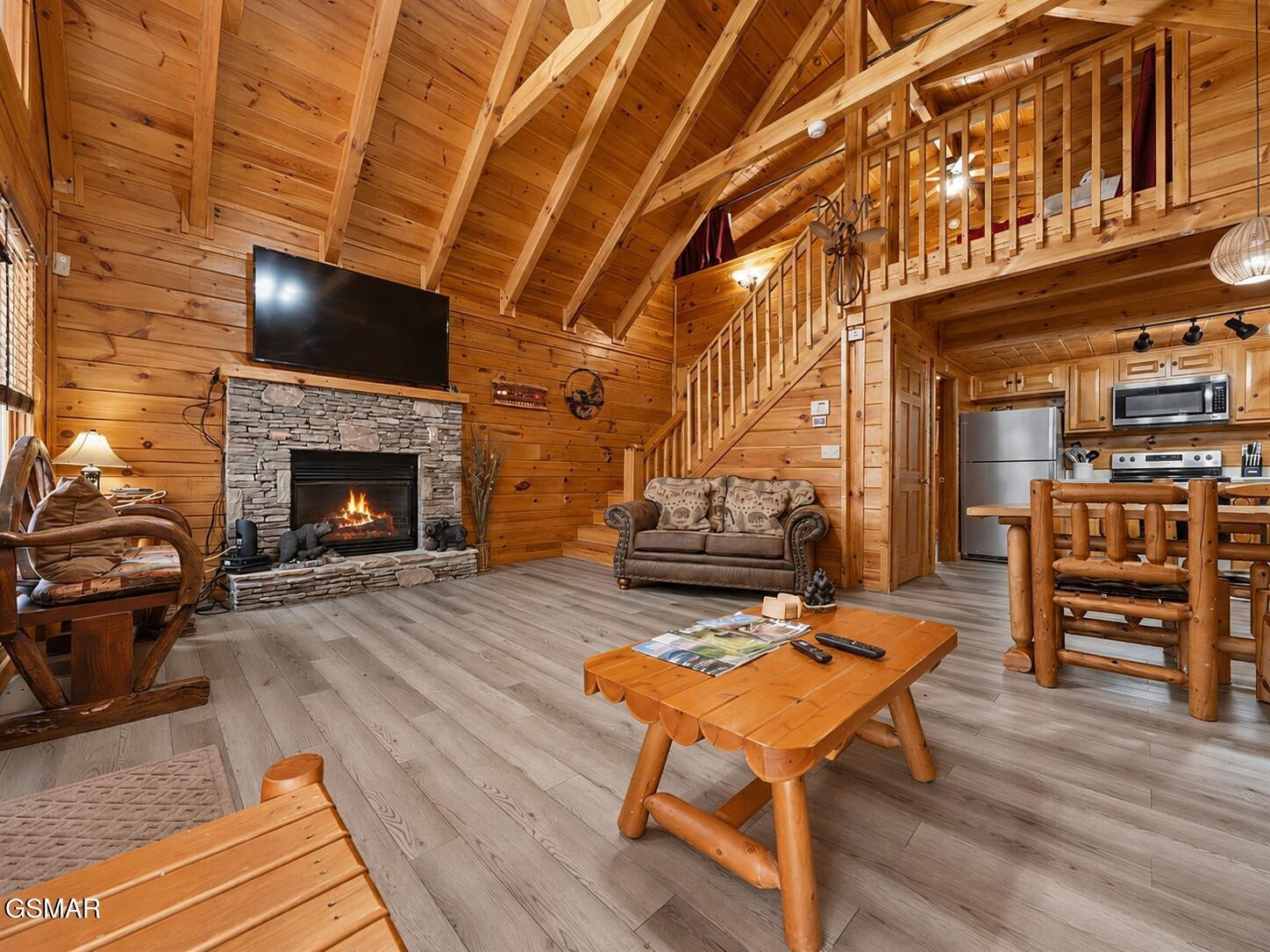 Rustic cabin living room with stone fireplace, vaulted wood ceiling, loft stairs, and open kitchen.
