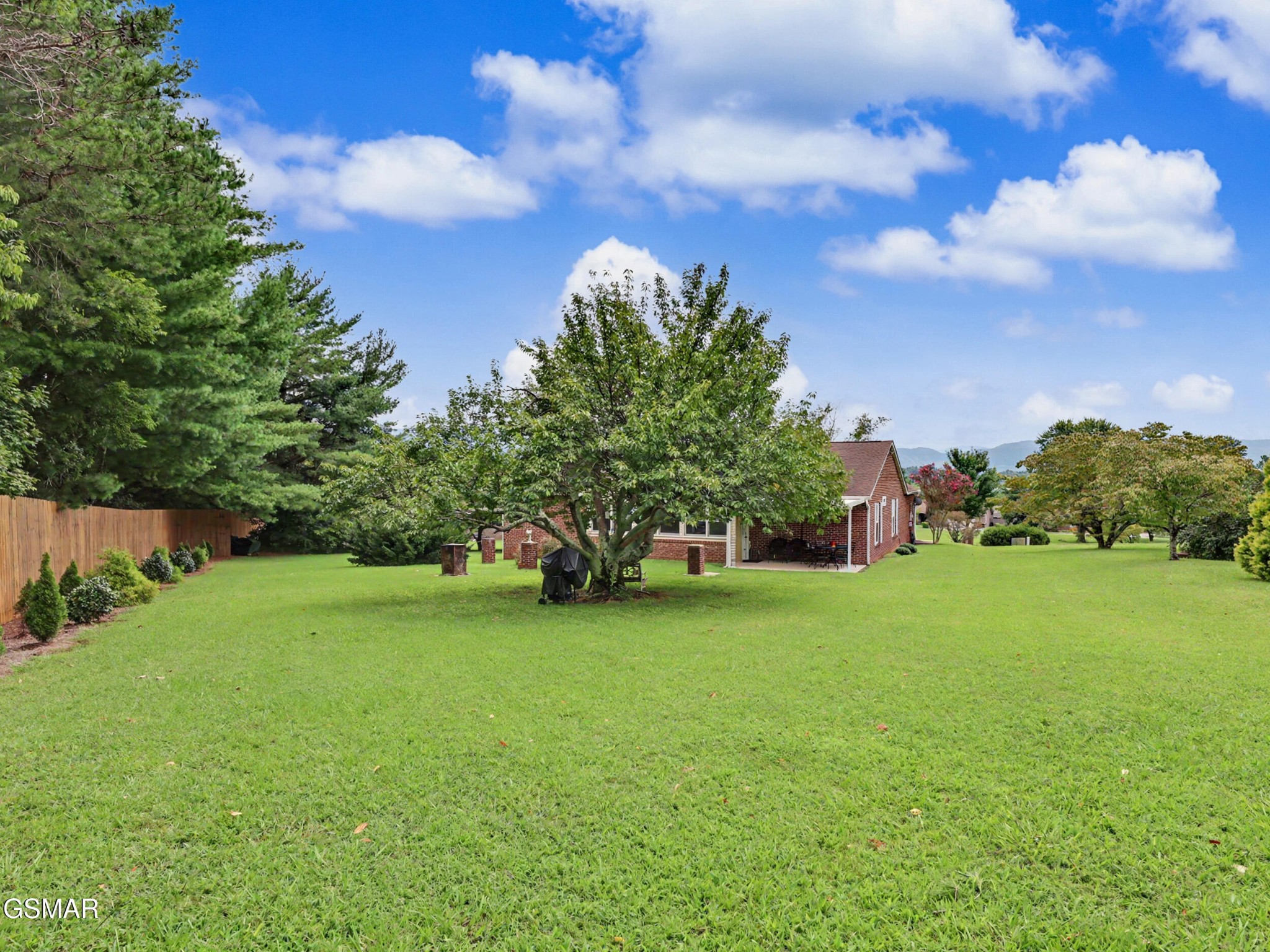 Brick home beyond a broad green backyard, with mature trees, a covered patio, and distant mountains.