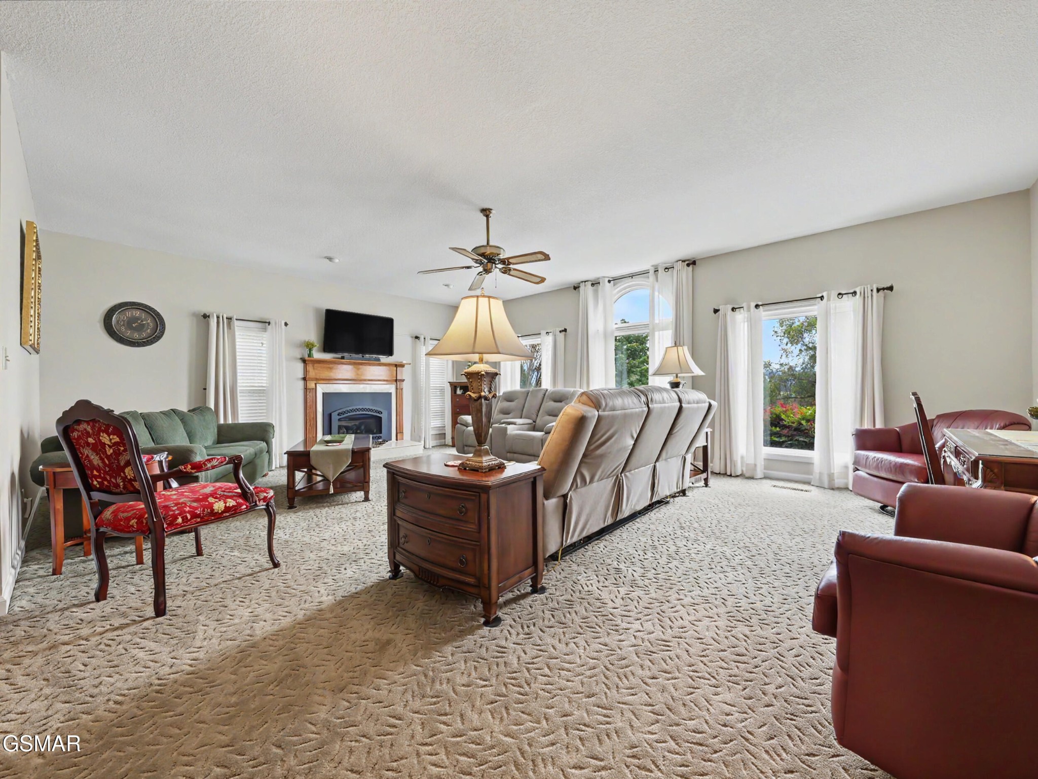 Furnished living room with a fireplace, multiple seating areas, and large windows overlooking greenery.