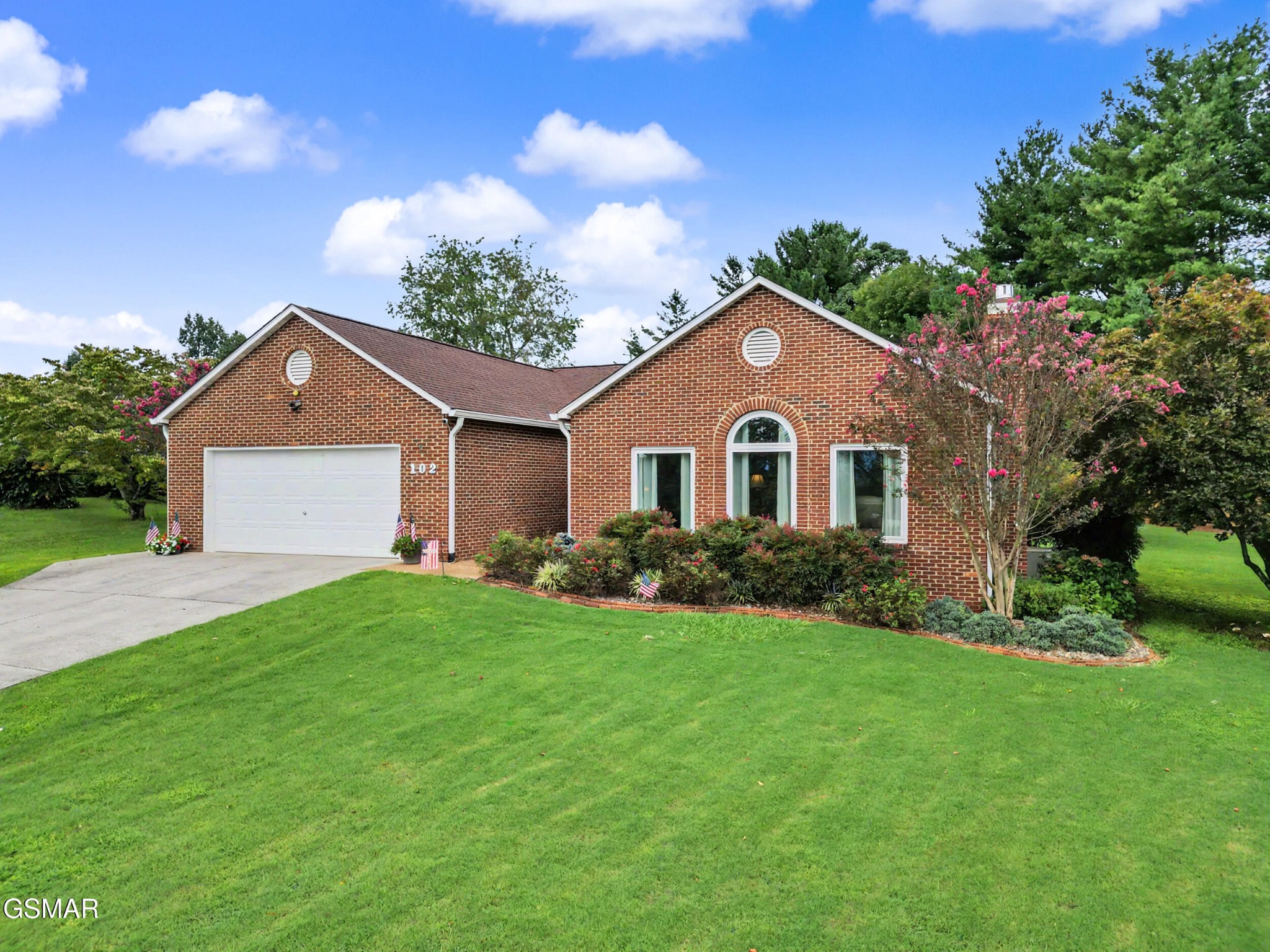 Red brick home with an attached garage, arched window, flowering landscaping, and broad lawn.