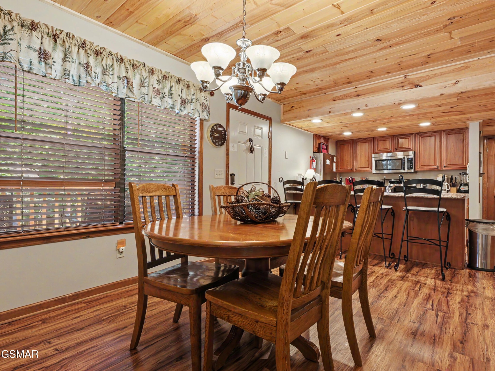 Dining area with a round wooden table, opening to a kitchen with a breakfast bar and wood-plank ceiling.