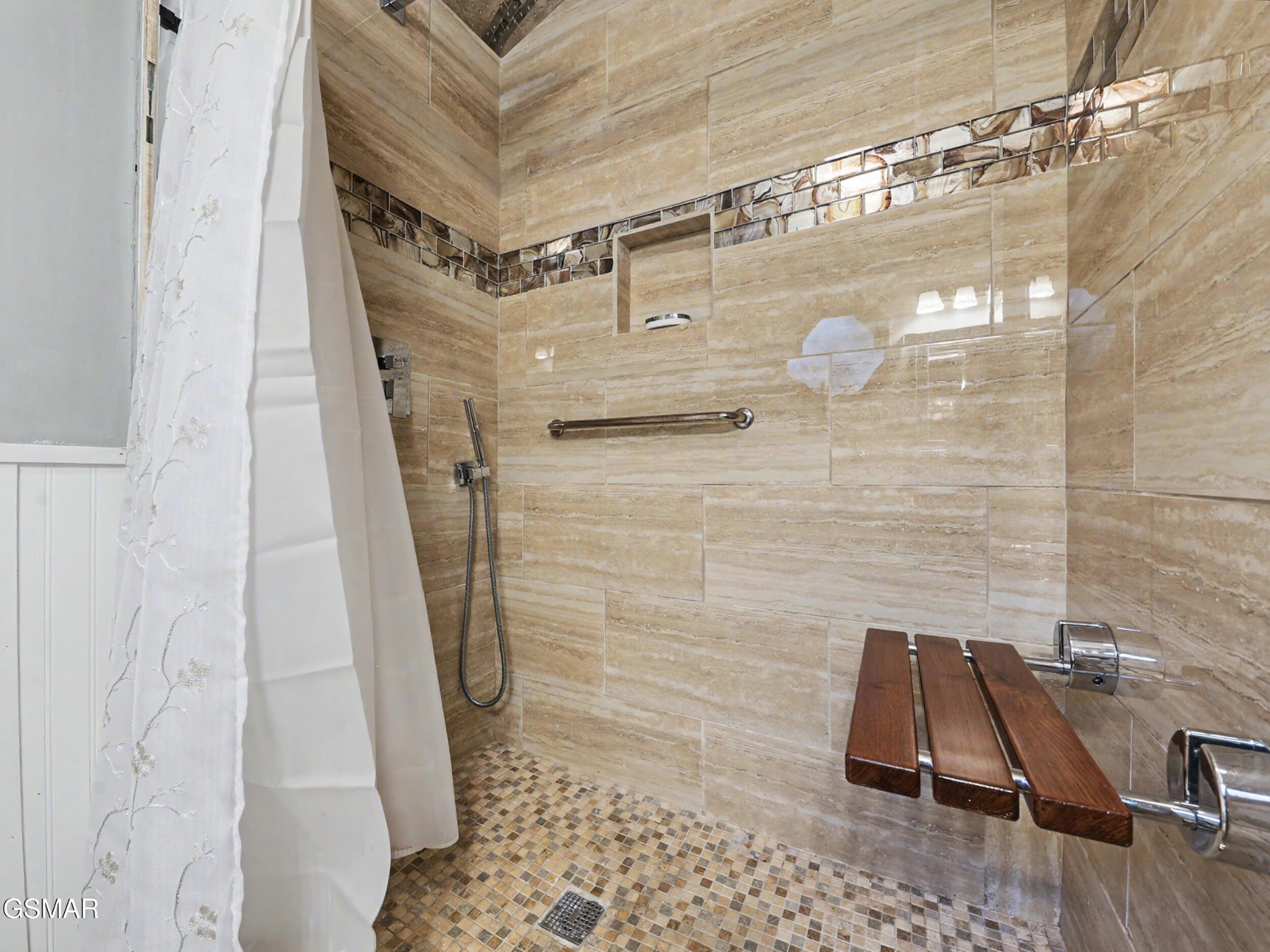 Walk-in shower with tan tile, mosaic floor, grab bar, and fold-down wooden seat.