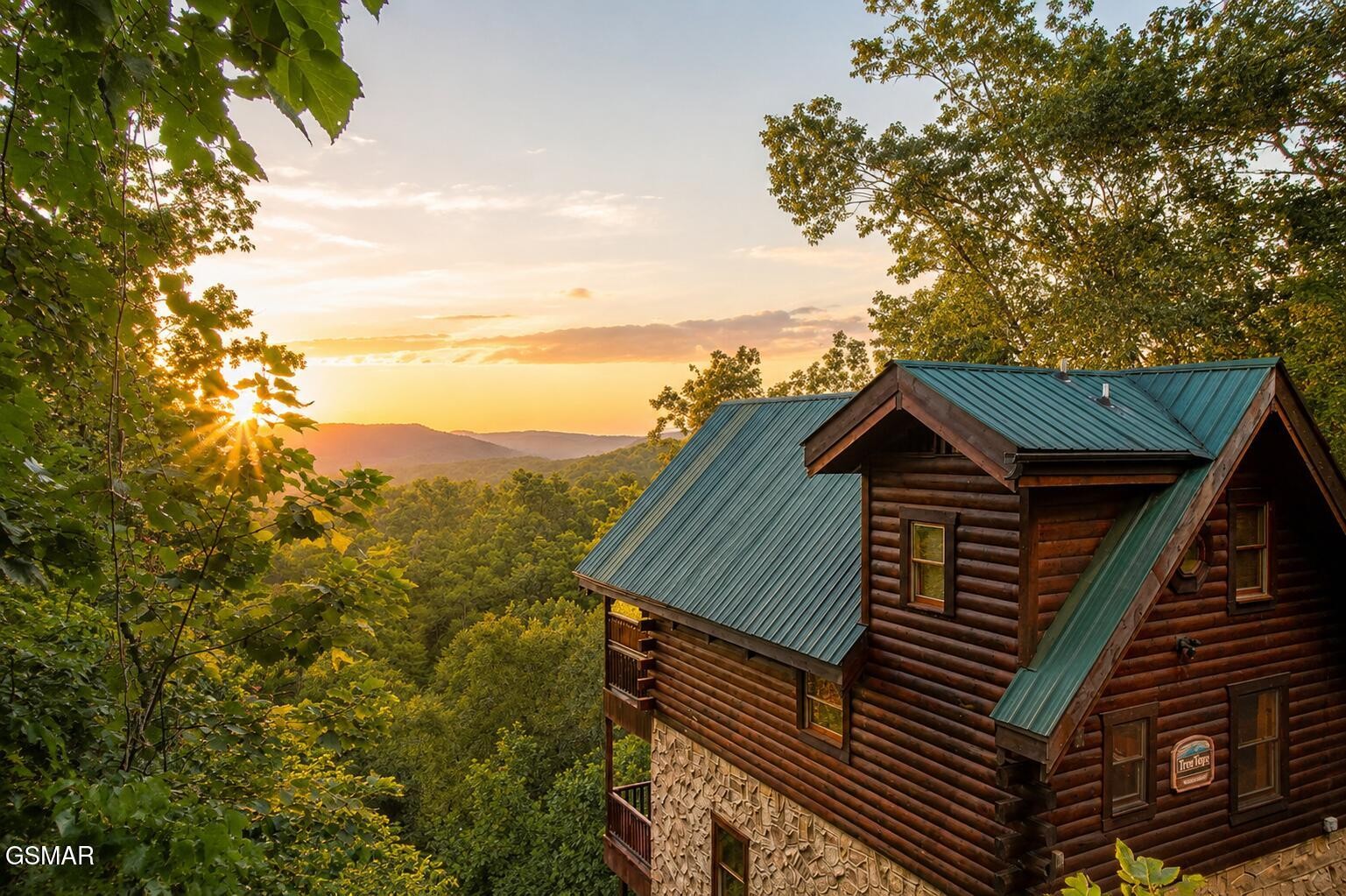 Log cabin with green metal roofs overlooking wooded mountain ridges at sunset.
