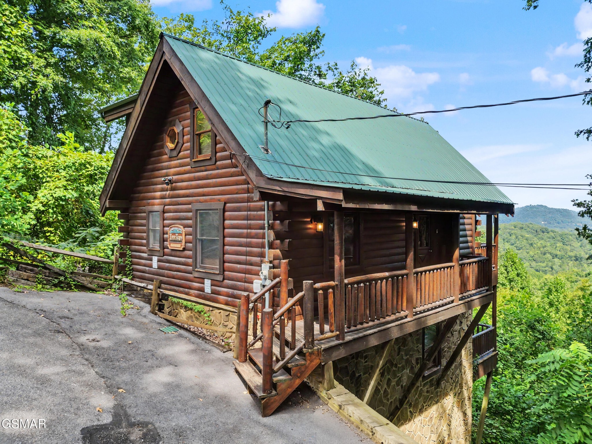 Log cabin with a green metal roof, raised covered porch, and wooded hills beyond.