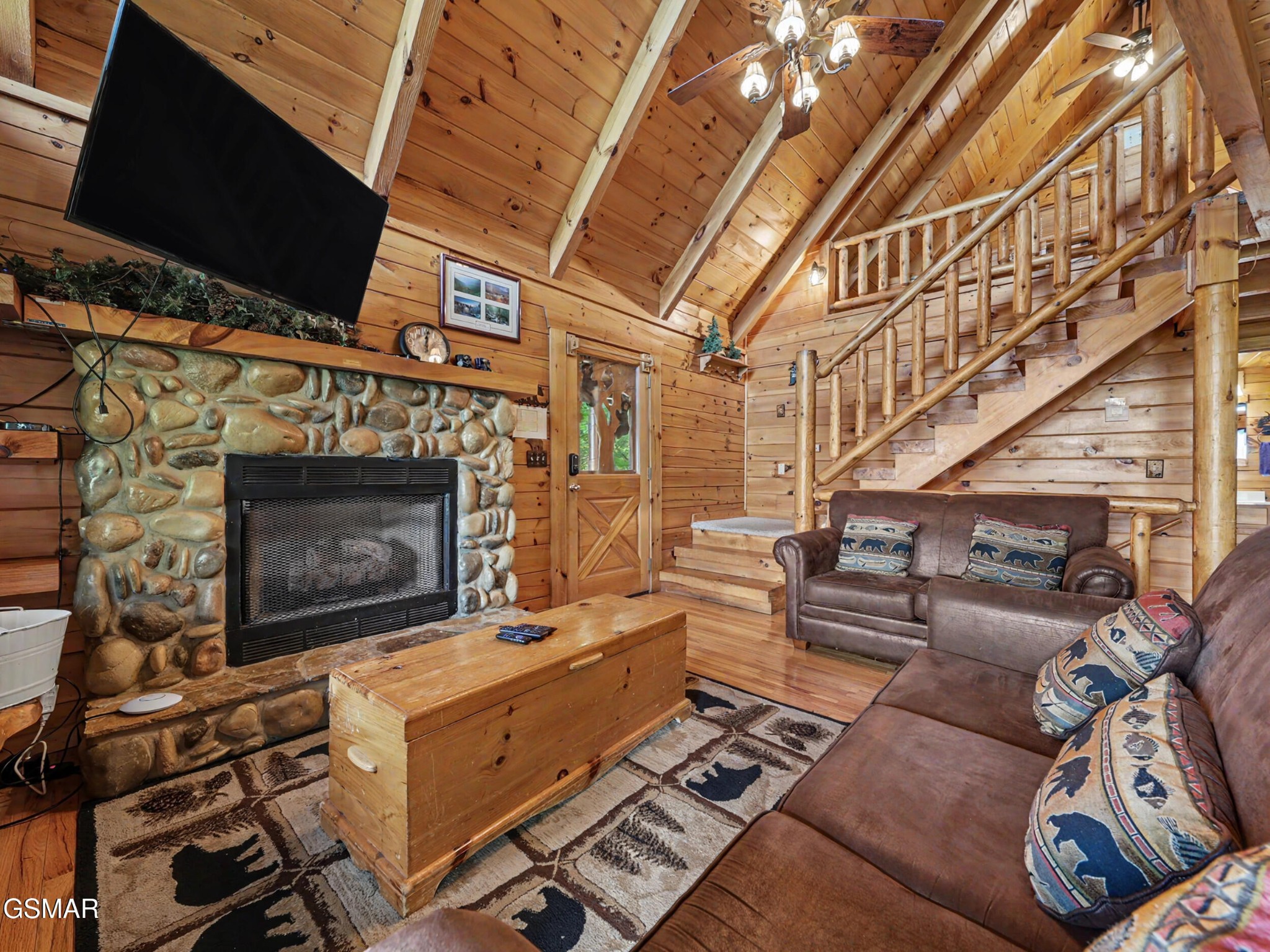 Cabin living room with a stone fireplace, vaulted wood ceiling, and log staircase.