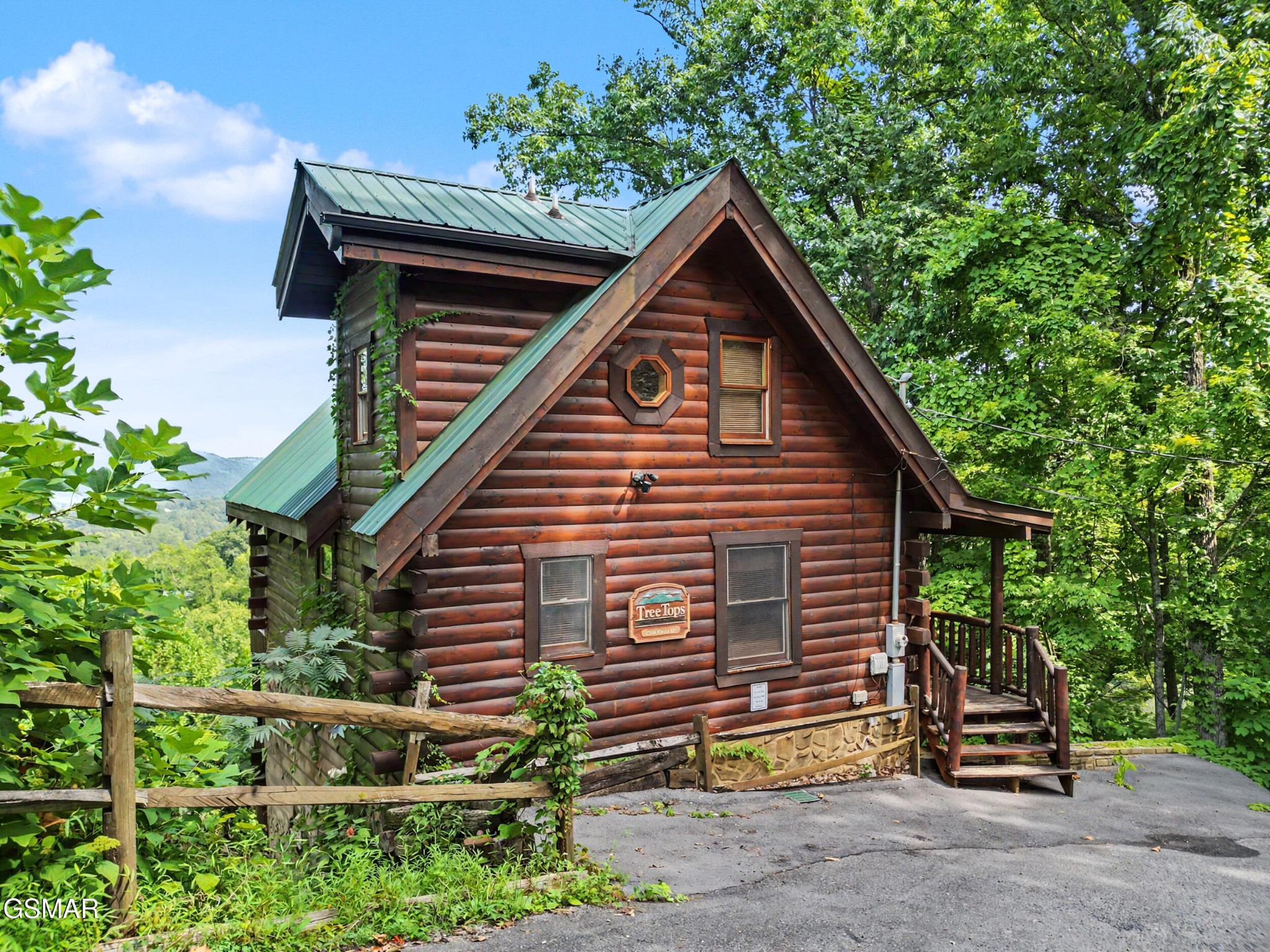 Two-story log cabin with a green metal roof, raised entry porch, and wooded mountain setting.