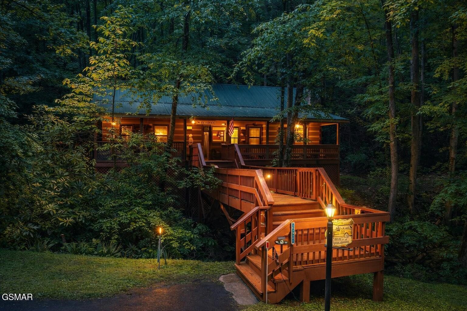 Wooded log cabin at dusk with a metal roof, illuminated deck, and long wooden staircase.