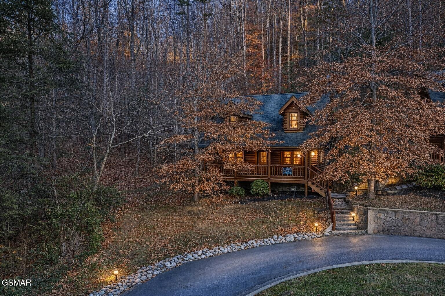 Log cabin among autumn trees at dusk, with a warmly lit porch and stone steps.