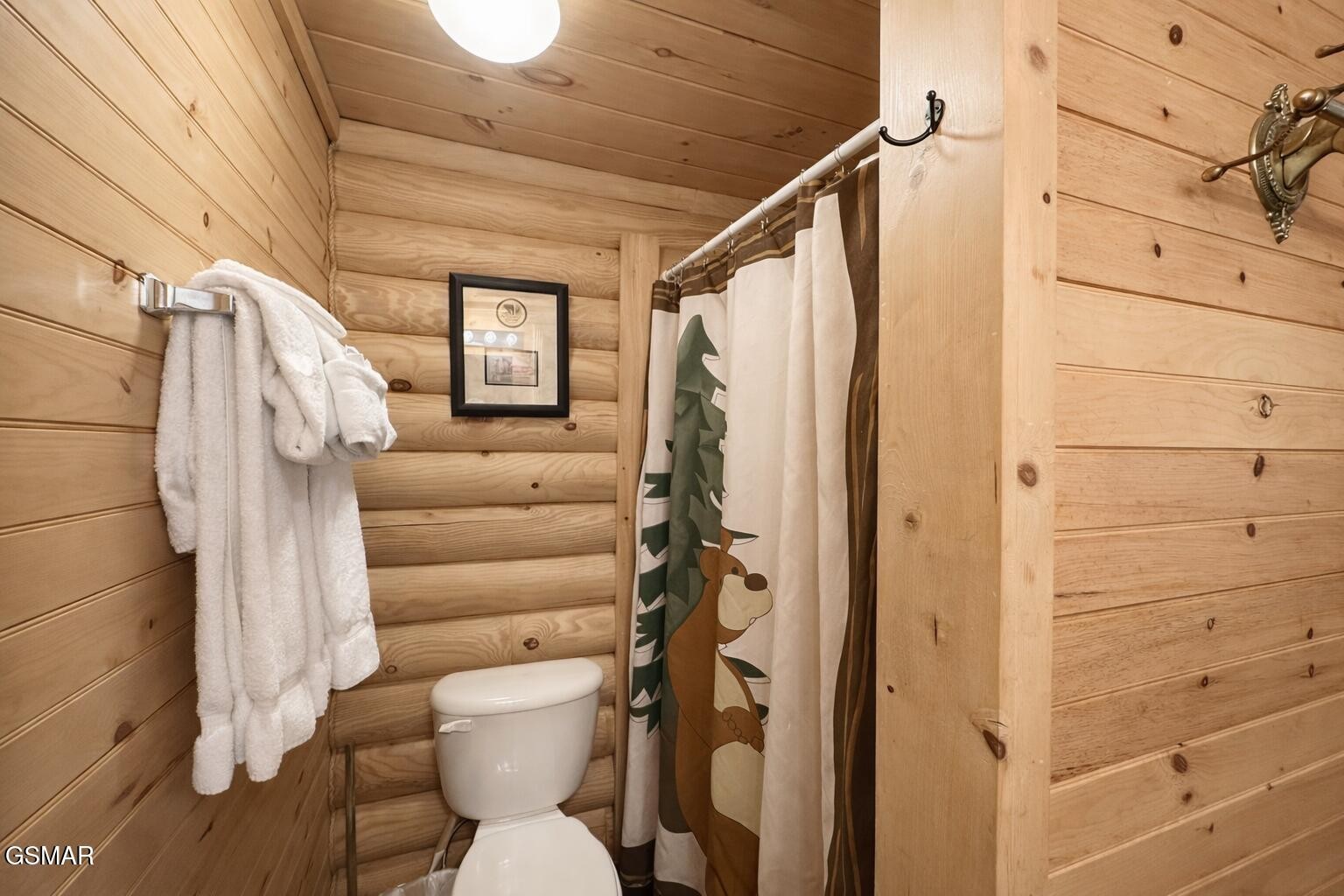 Rustic log cabin bathroom with a deer-print shower curtain, white toilet, and towels on a wall rack.