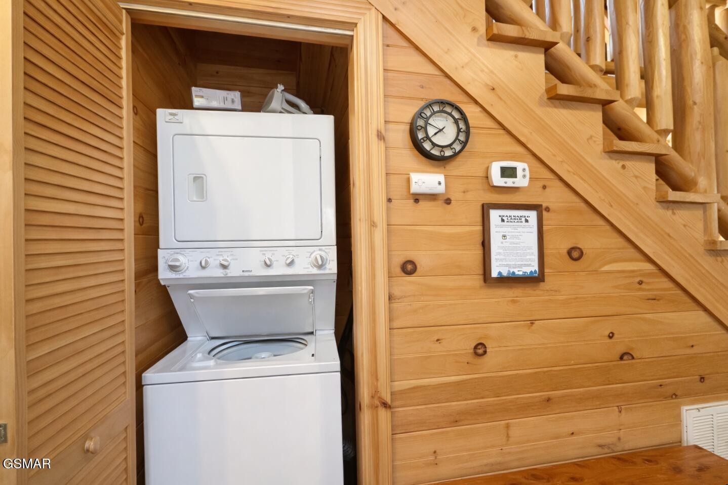 Stacked washer and dryer tucked beneath a wooden staircase in a wood-paneled cabin.