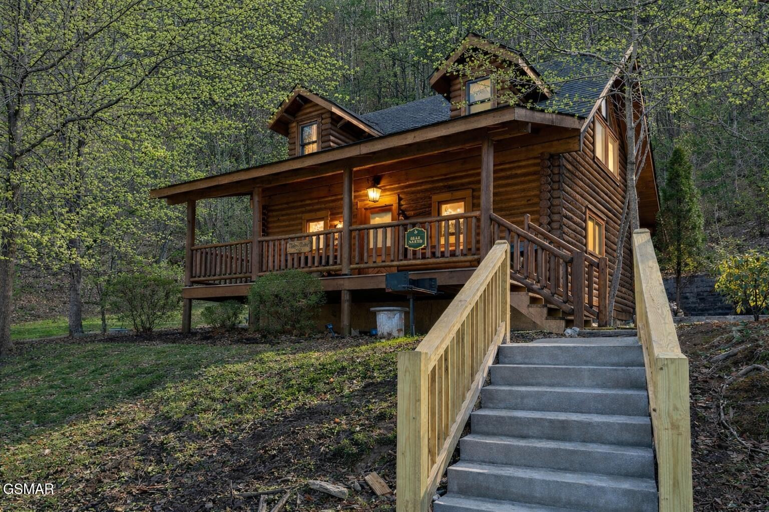 Log cabin with a covered porch and concrete steps, nestled among budding trees.