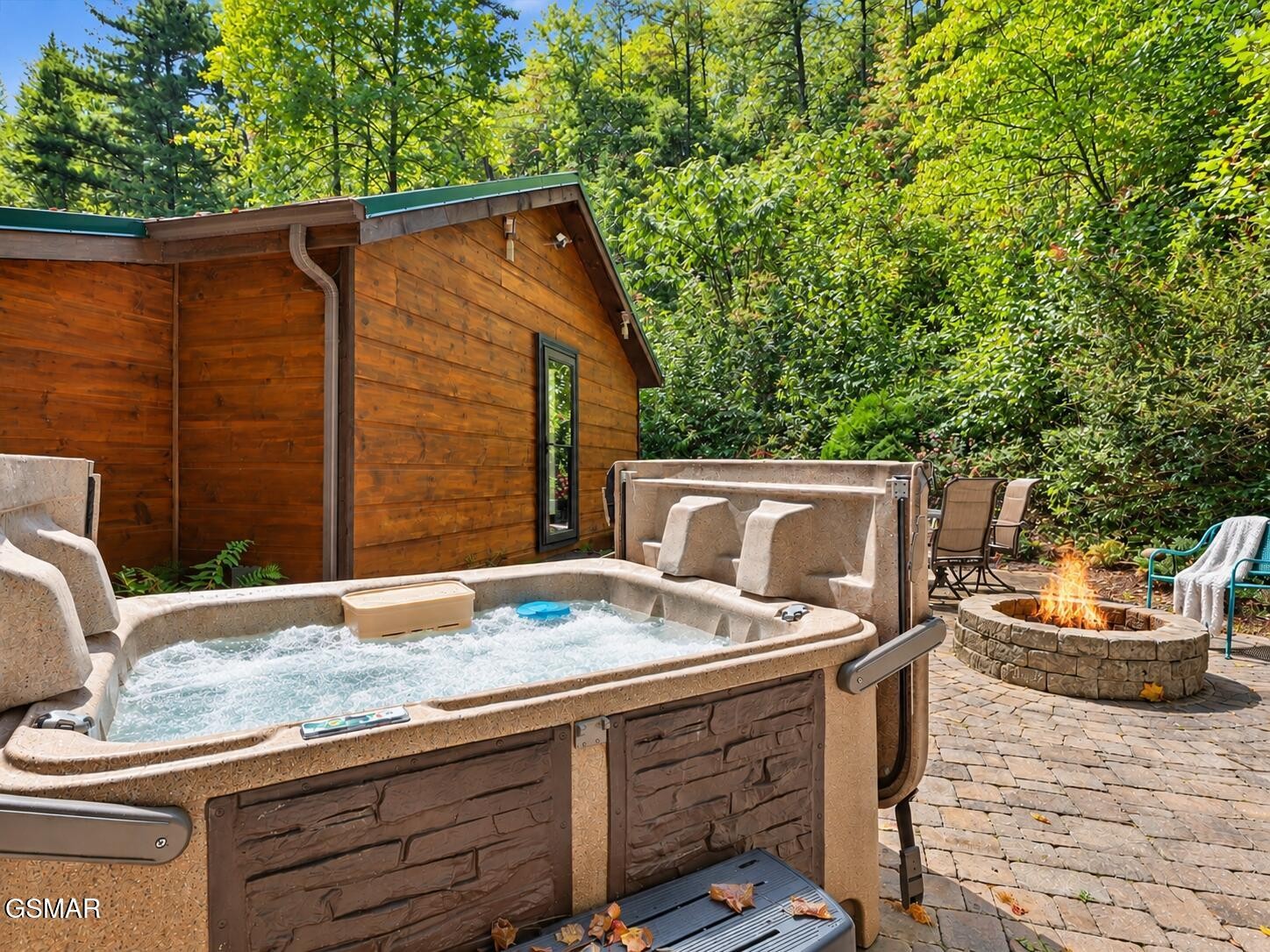 Outdoor hot tub and stone fire pit beside a wood-sided cabin, surrounded by dense trees.