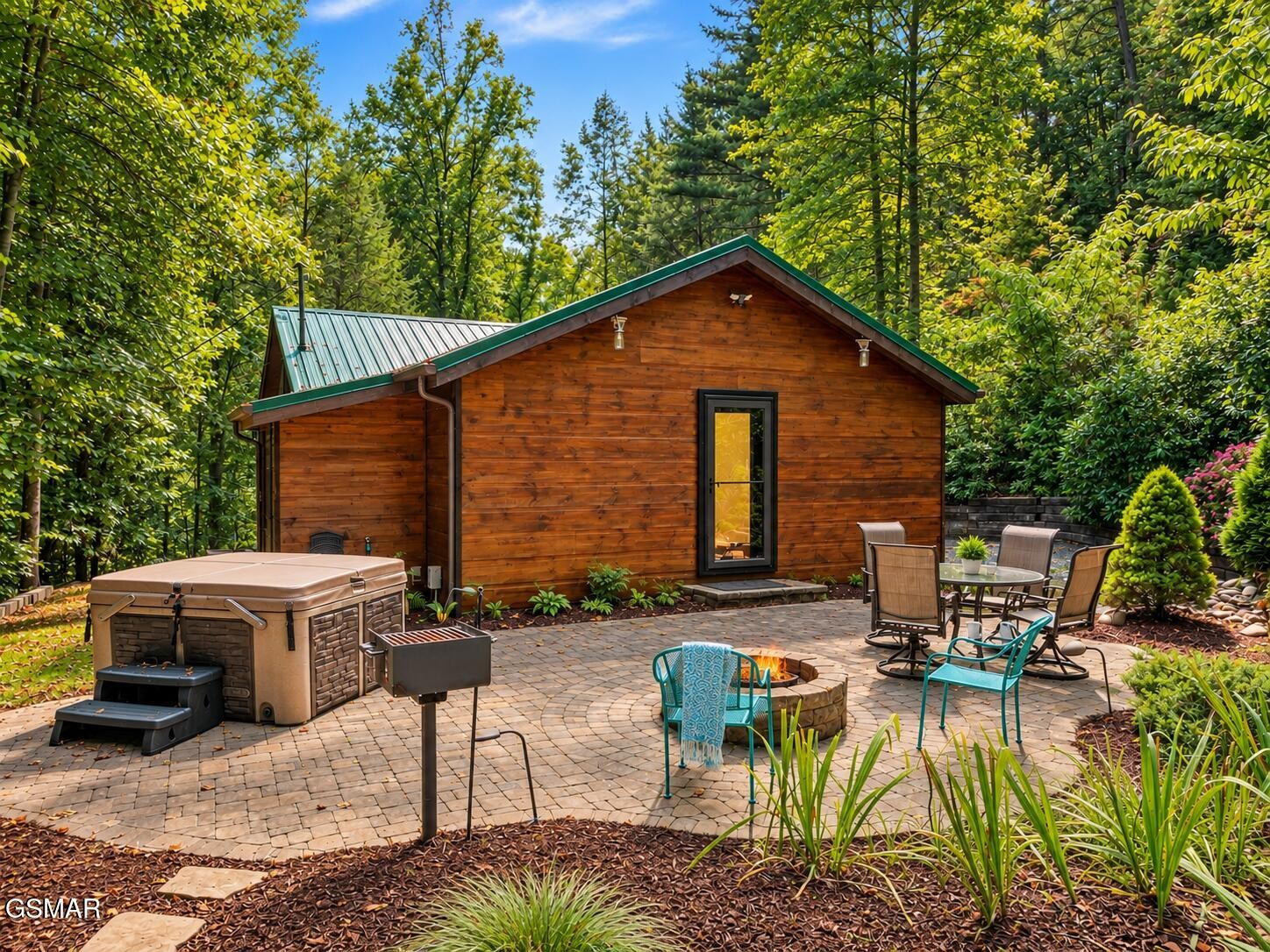 Wood-sided cabin with a green metal roof, hot tub, and fire pit on a wooded patio.