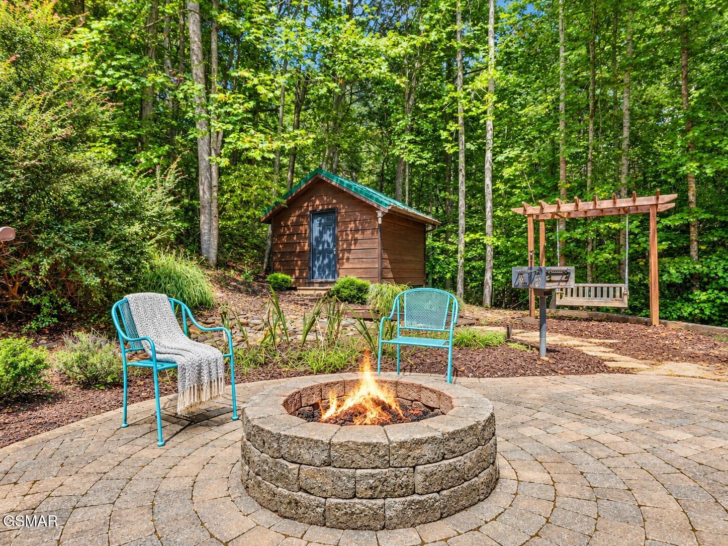 Wooded patio with a stone fire pit, turquoise chairs, shed, and pergola swing.