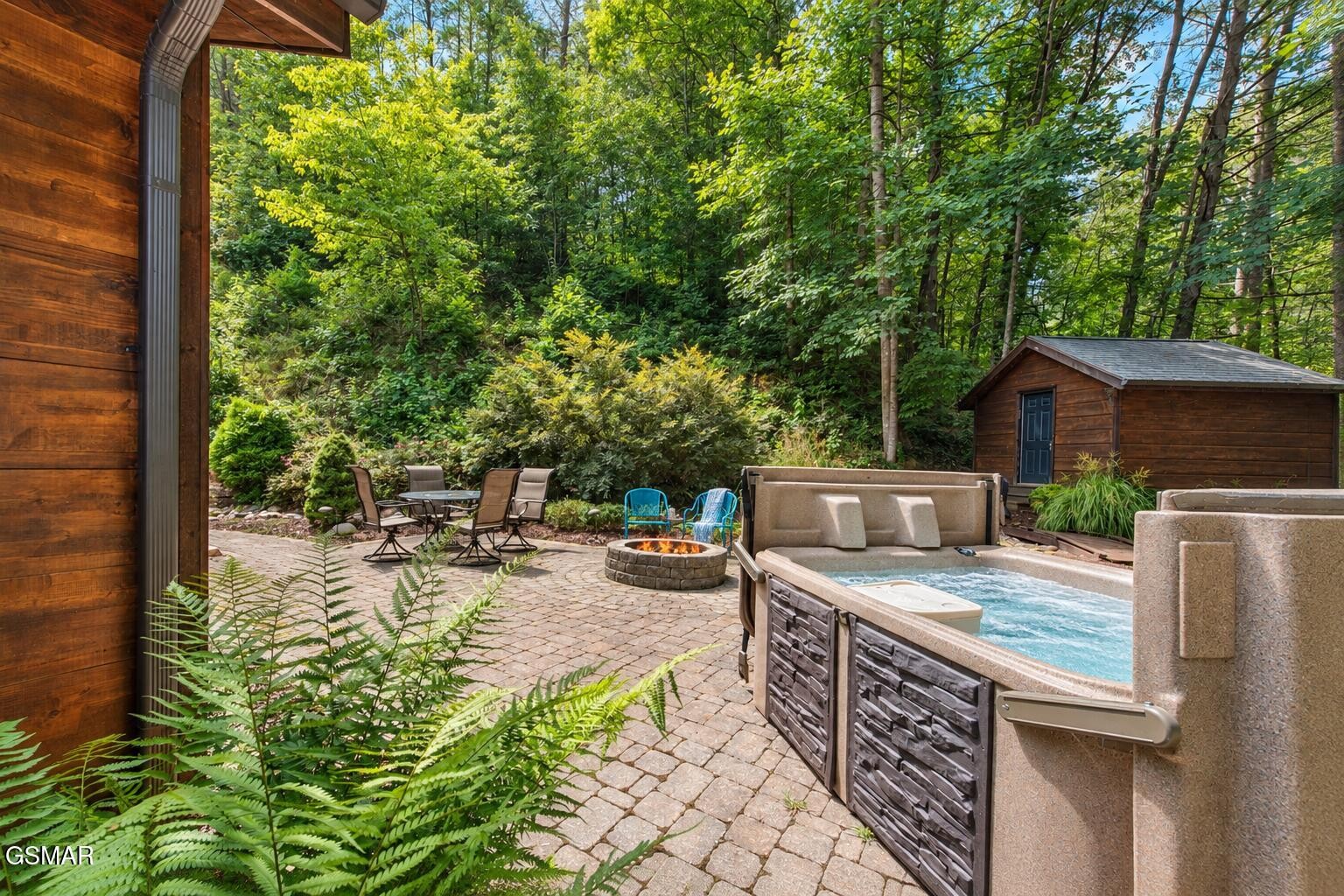 Wooded backyard patio with a bubbling hot tub, stone fire pit, dining set, and wooden shed.