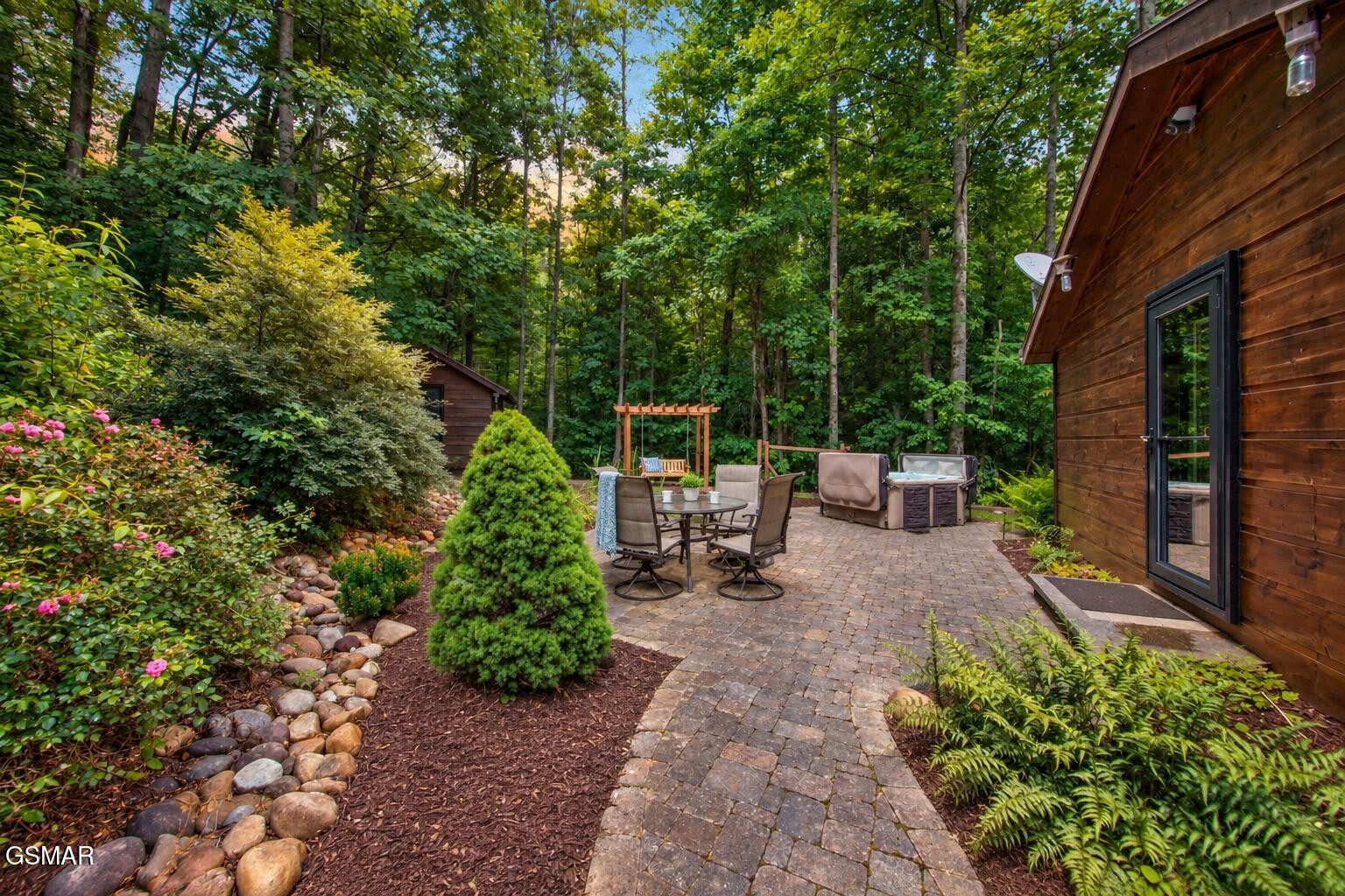 Cabin patio with paver walkway, outdoor dining set, lounge seating, and surrounding woods.